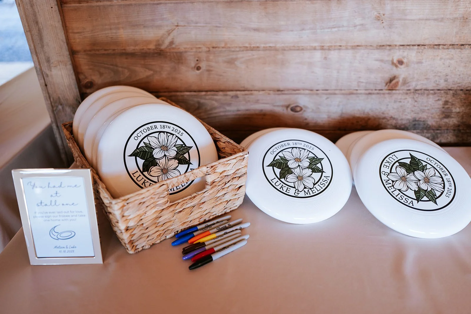 White frisbees with floral design, personalized with the names Luke and Melissa and the date October 18th, 2025, arranged on a table with pens and a sign.