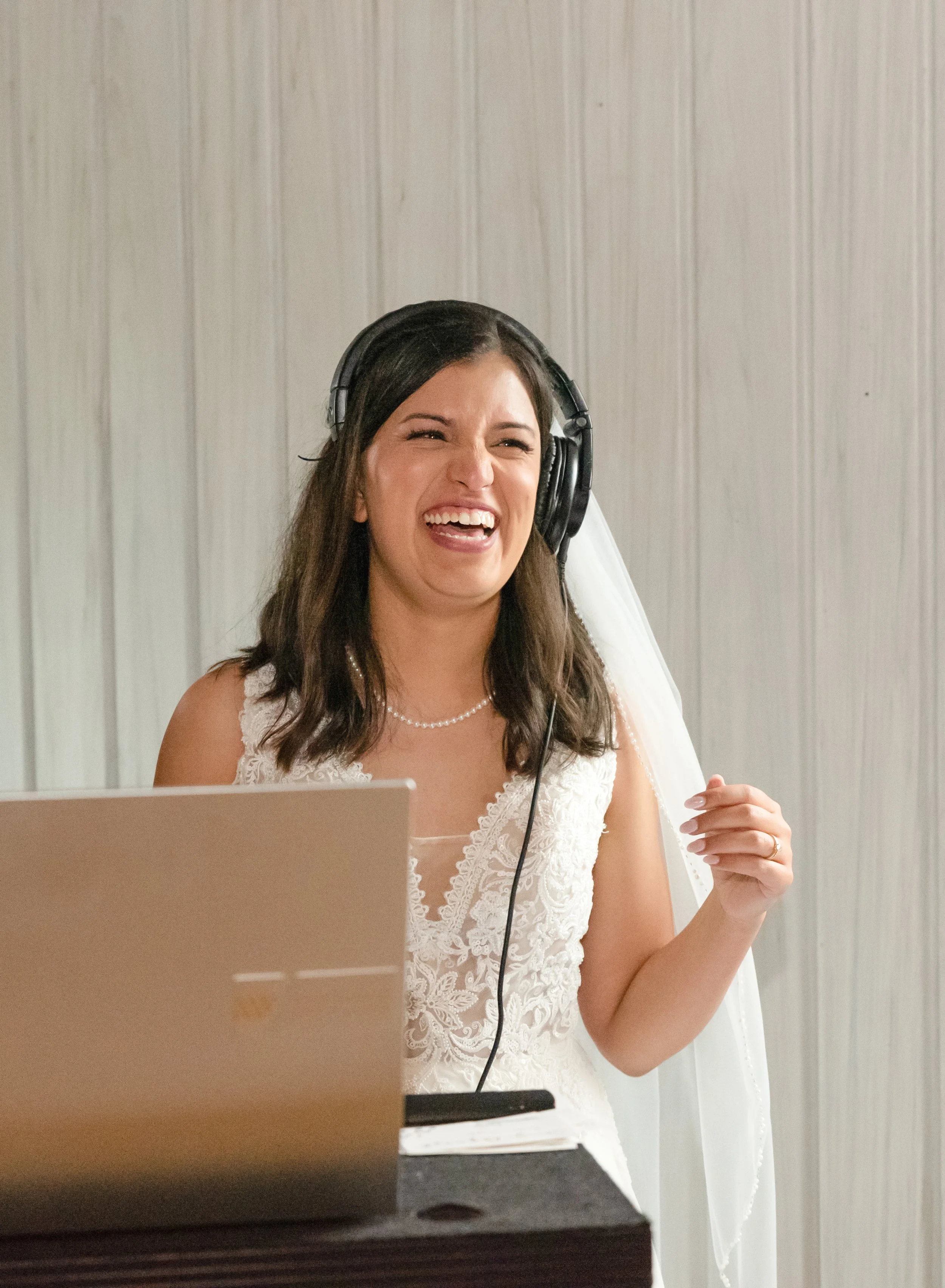A woman in a wedding dress wearing headphones, smiling and singing into a microphone in front of a laptop.