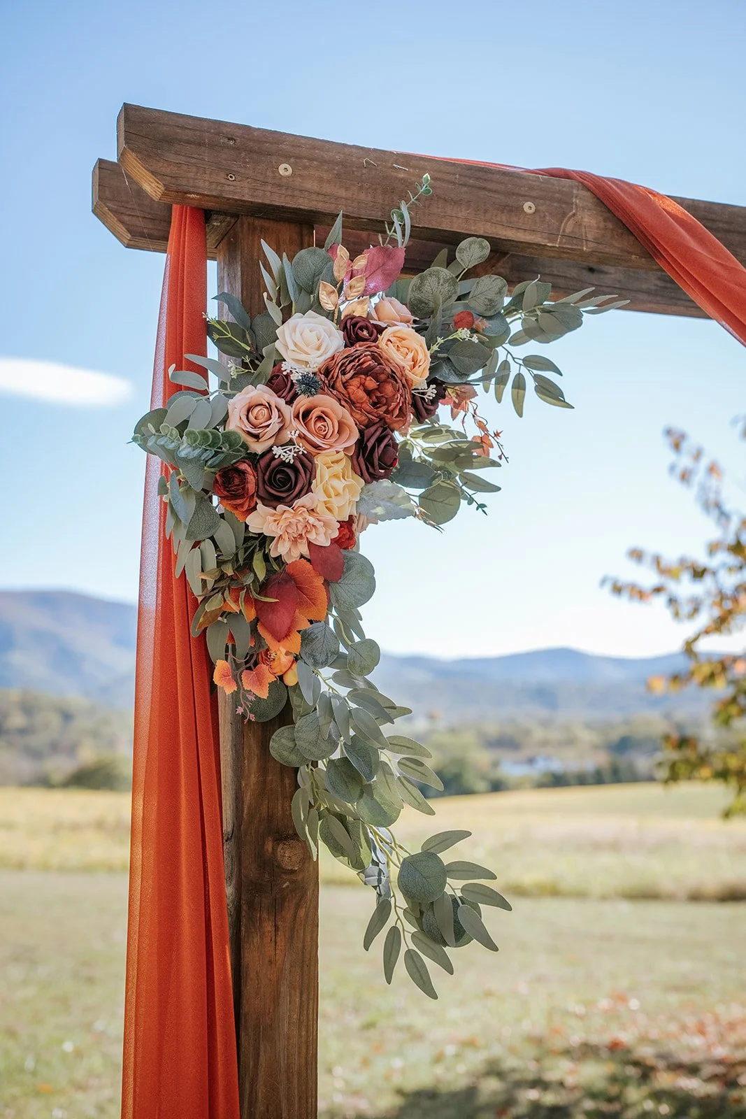 Floral arrangement with roses and eucalyptus leaves on a wooden outdoor structure, orange fabric draped on a clear day with hills in the background.