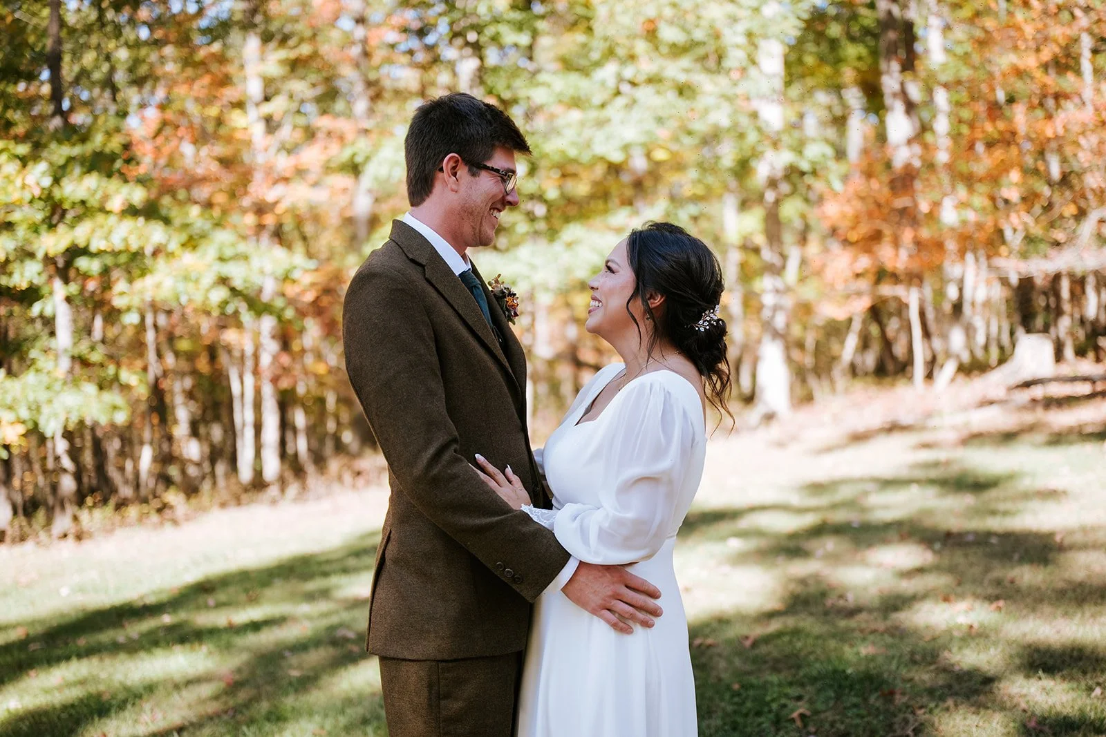 A couple dressed in wedding attire standing outdoors amidst fall foliage, gazing into each other's eyes and smiling.