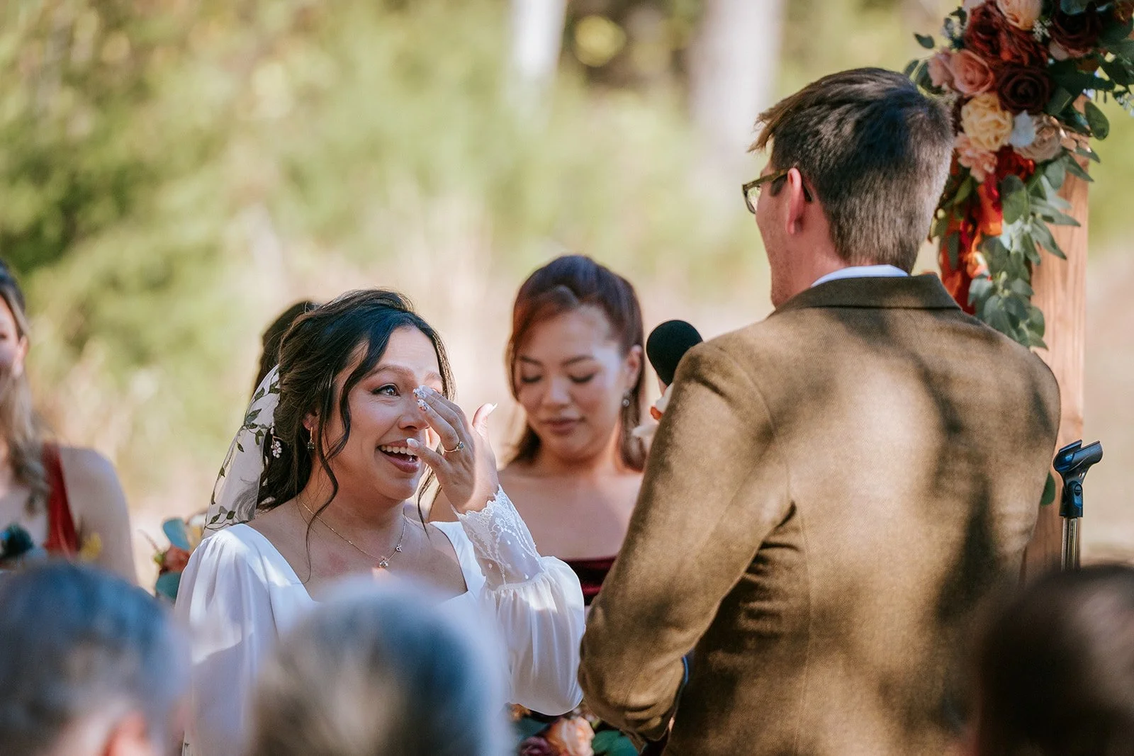 A woman in a white dress wiping away tears during a wedding ceremony, with a man in a brown suit officiating, outdoors with a floral arch in the background.