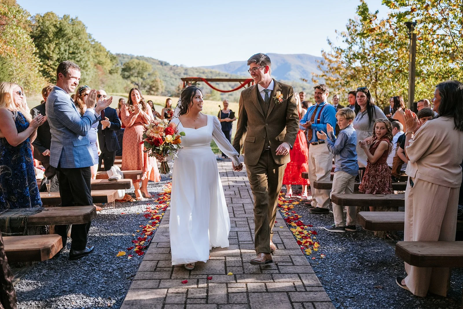 A bride and groom walking hand in hand down the aisle at an outdoor wedding ceremony, surrounded by guests on either side, with a scenic mountain landscape in the background on a sunny day.