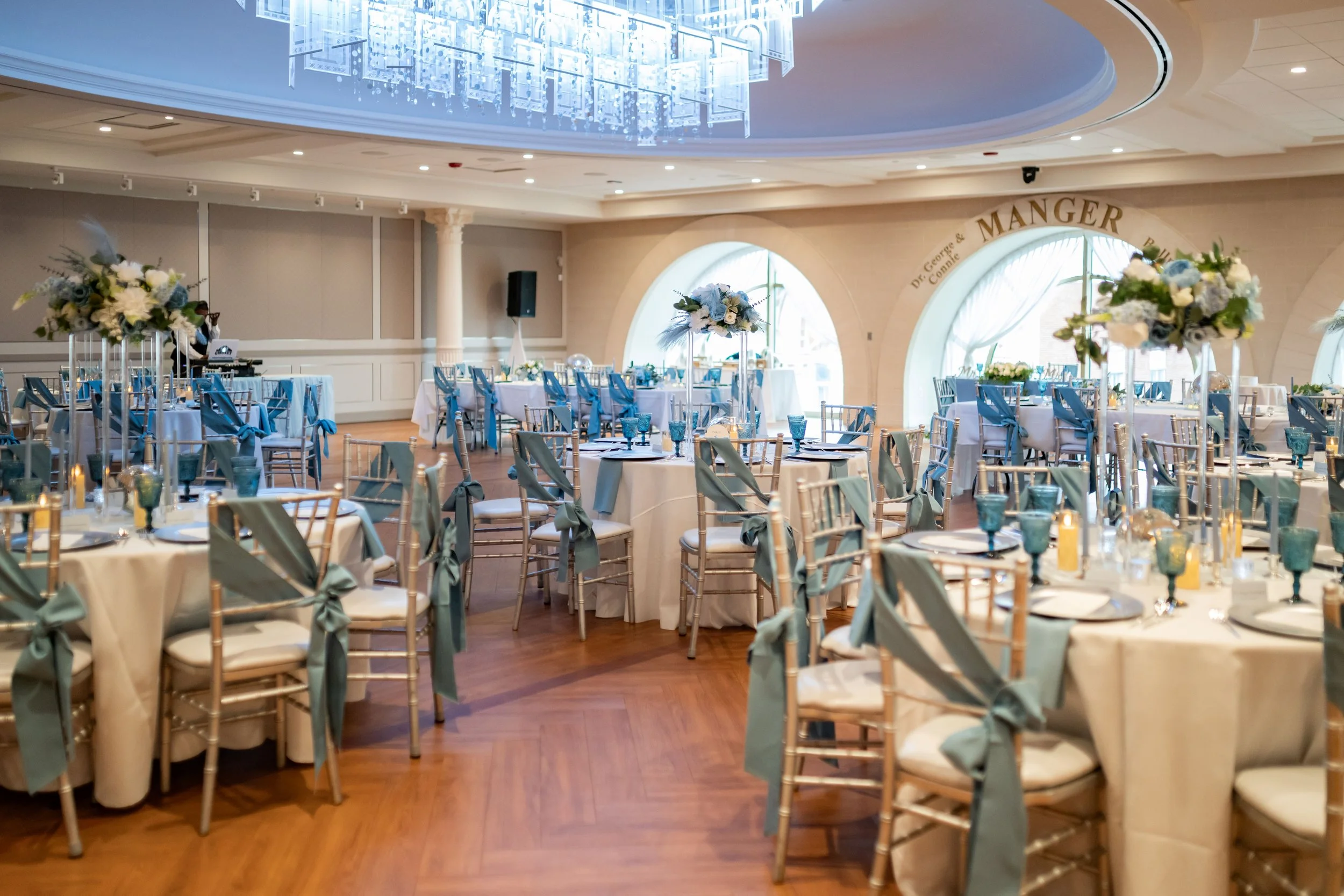 The ballroom at the Maryland Theater, l decorated for a wedding reception with round tables, blue and white floral centerpieces, gold chairs with blue ribbons, and a chandelier overhead.
