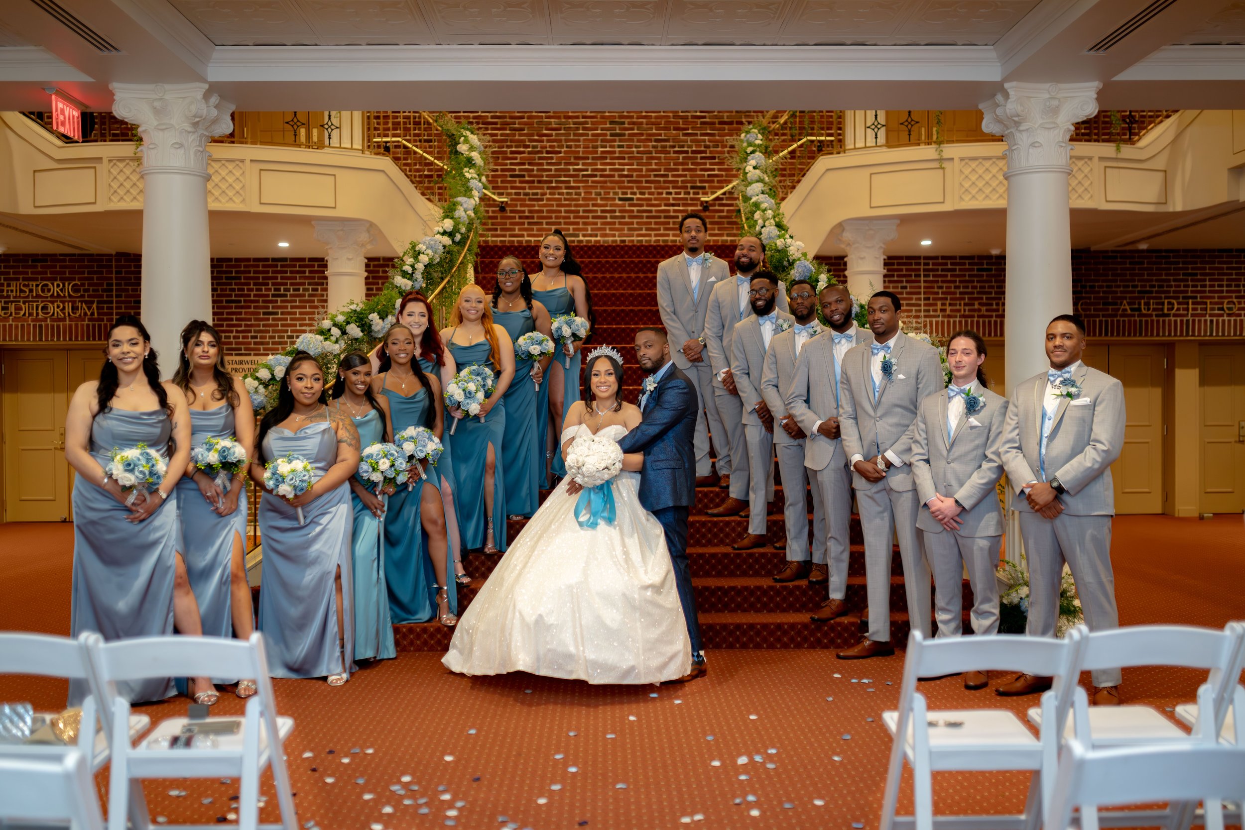 Group photo of a wedding party on a staircase with floral decorations. The bride and groom are in the center, with bridesmaids in blue dresses on the left and groomsmen in light gray suits on the right.