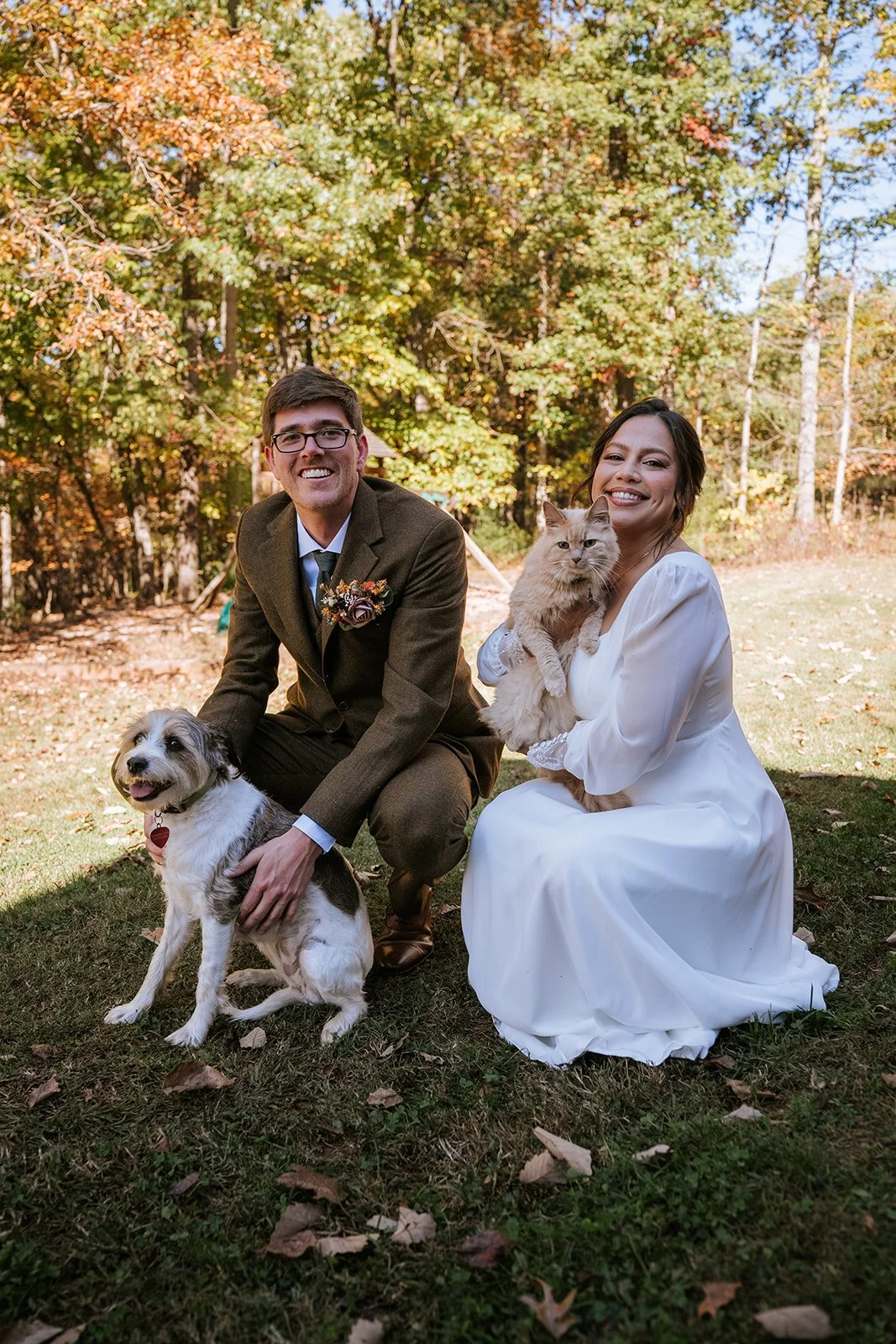 A couple dressed in wedding attire, with the groom in a suit and the bride in a white gown, outdoors with autumn trees in the background. They are holding two cats and accompanied by a dog.