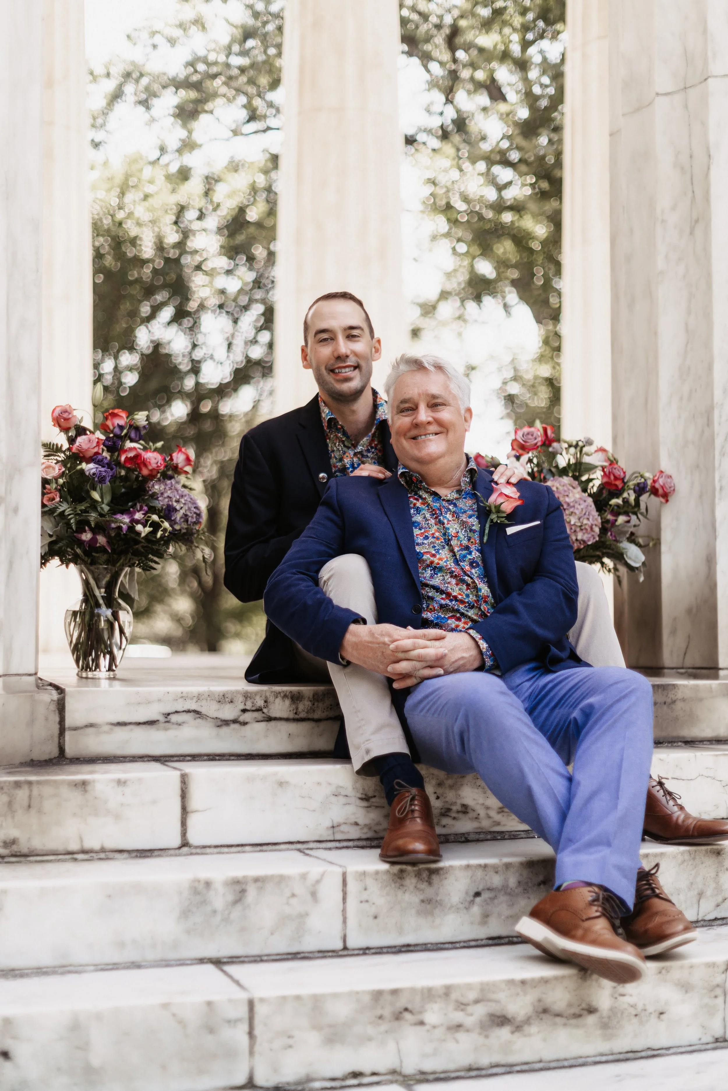 Two men sitting on marble stairs in front of white pillars, each wearing a floral shirt and blazer. Bouquet of colorful flowers arranged on either side.