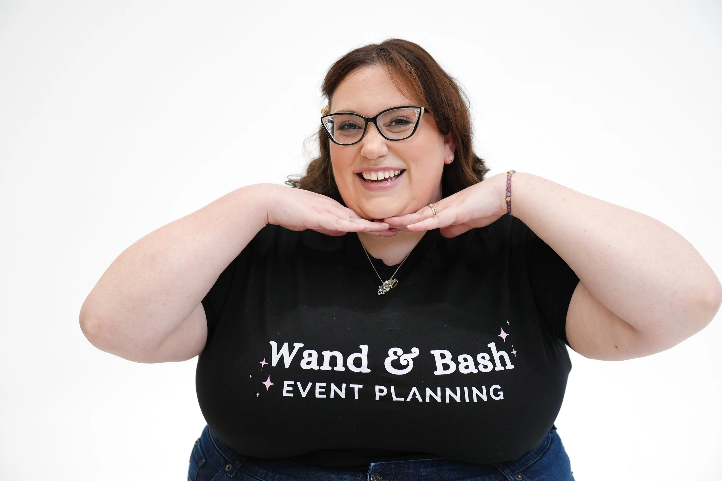 Woman with red hair, glasses, and a big smile posing with her hands under her chin against a white background. She is wearing a black t-shirt with the text 'Wand & Bash EVENT PLANNING' and is dressed casually with a purple beaded bracelet and a neckl
