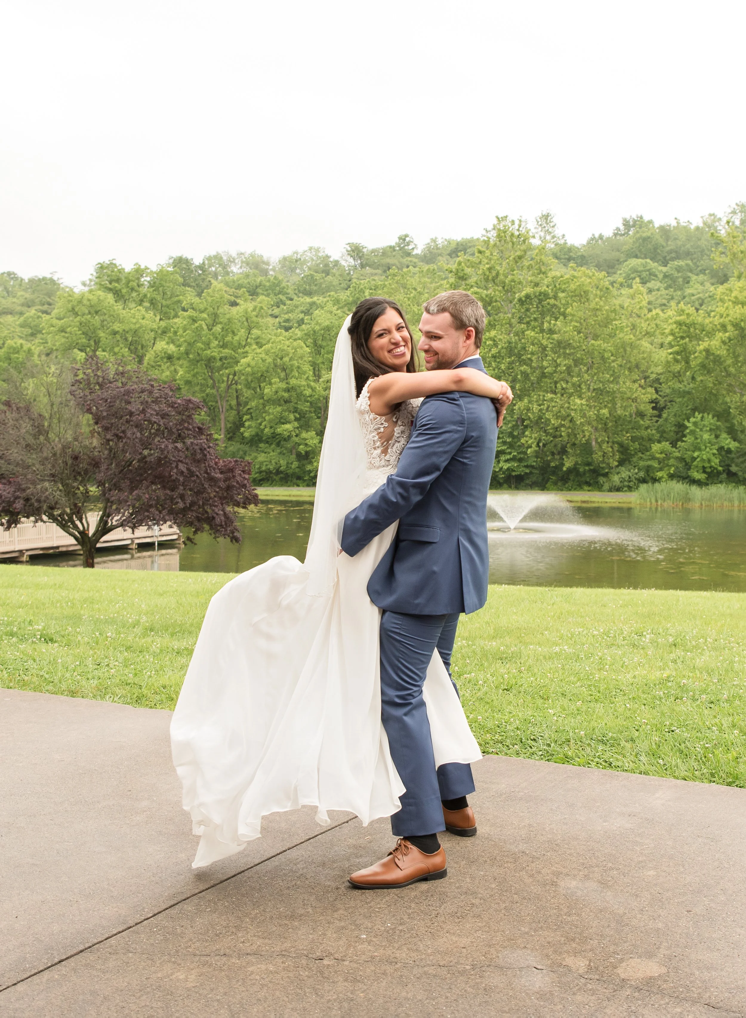 A couple in wedding attire dancing outdoors near a pond with a fountain, surrounded by green trees.