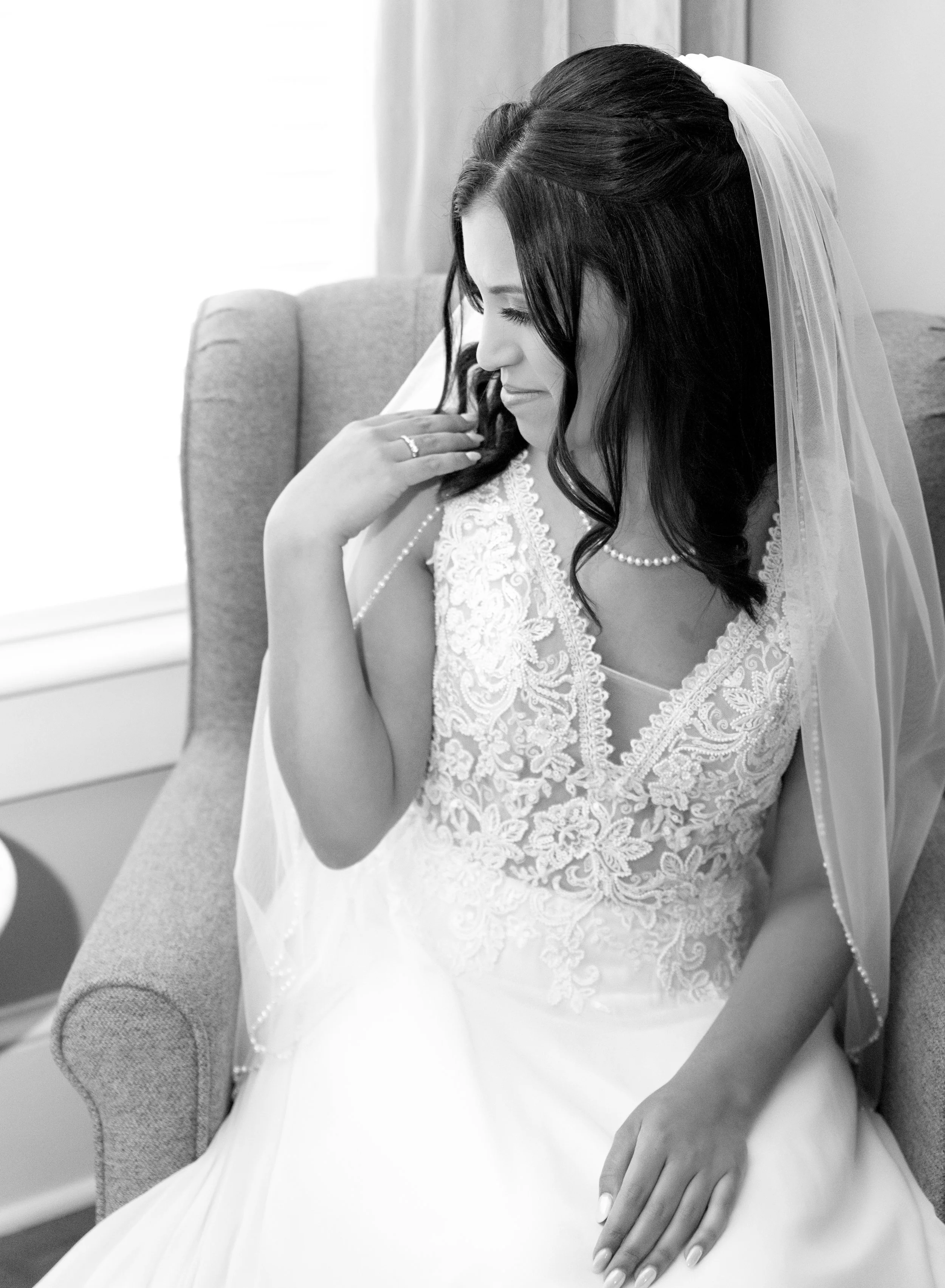 A bride sitting on a chair dressed in a lace wedding gown with a veil, touching her face gently, and smiling softly.
