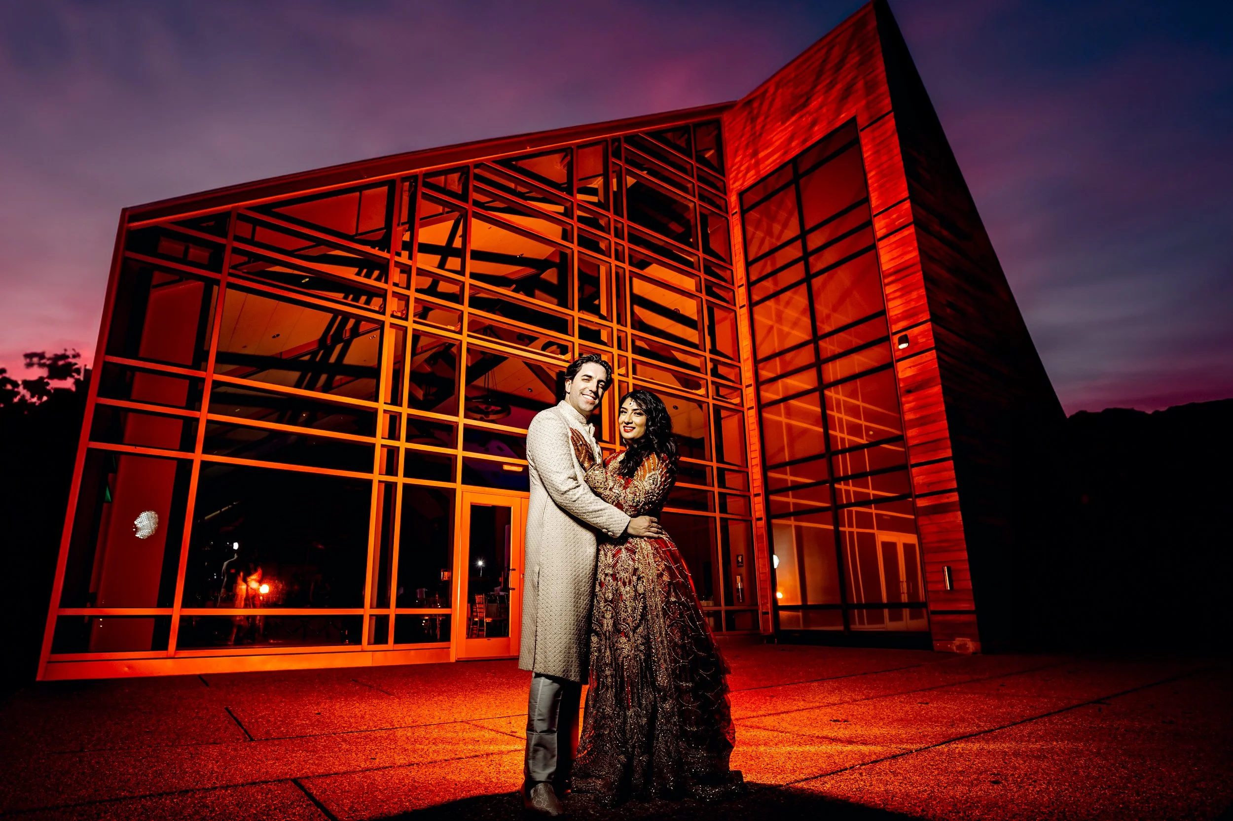 A couple dressed in traditional Indian attire, standing and embracing in front of a modern glass building with orange lighting during sunset.