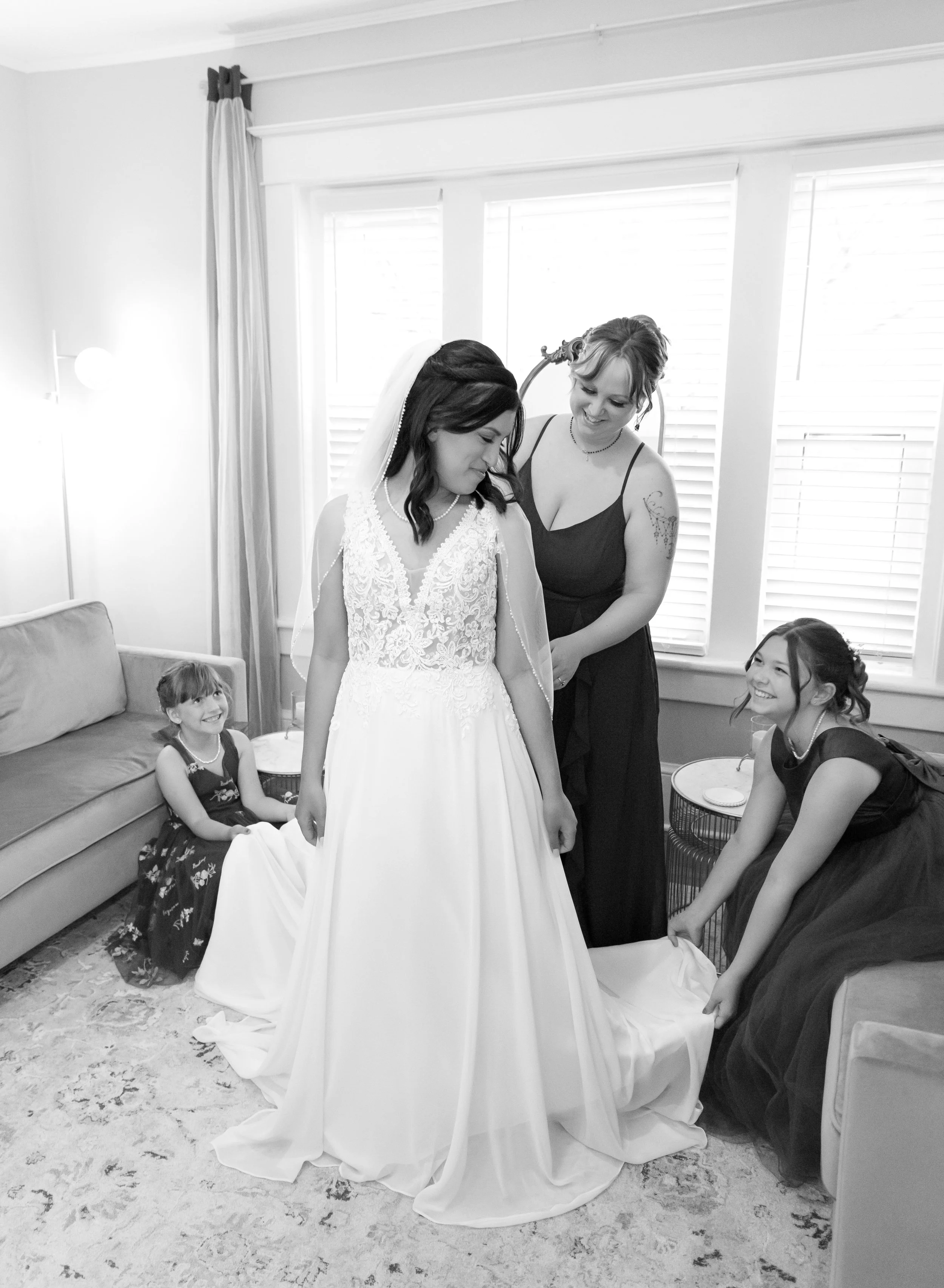 A bride in a wedding dress with an intricate lace bodice and a flowing skirt, standing in a room. Two women, one in a black dress and another in a sleeveless dress, are helping her with her dress. A young girl in a dark dress is sitting on a couch, s