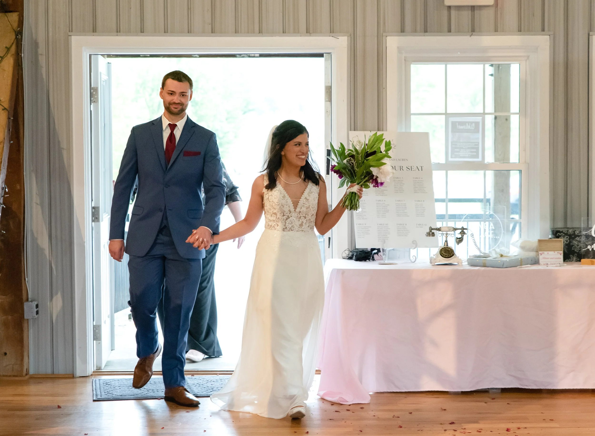 Bride and groom enter reception hand in hand. The bride holds a bouquet of flowers. They are in a rustic indoor setting with natural light.
