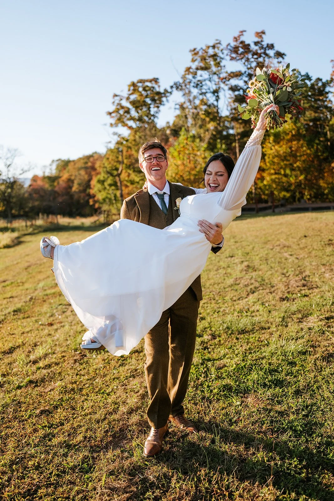 A couple, the groom in a suit and the bride in a white wedding dress, standing outdoors on a grassy field with autumn trees in the background. The groom is holding the bride, who is holding a bouquet, and they are both smiling joyfully.