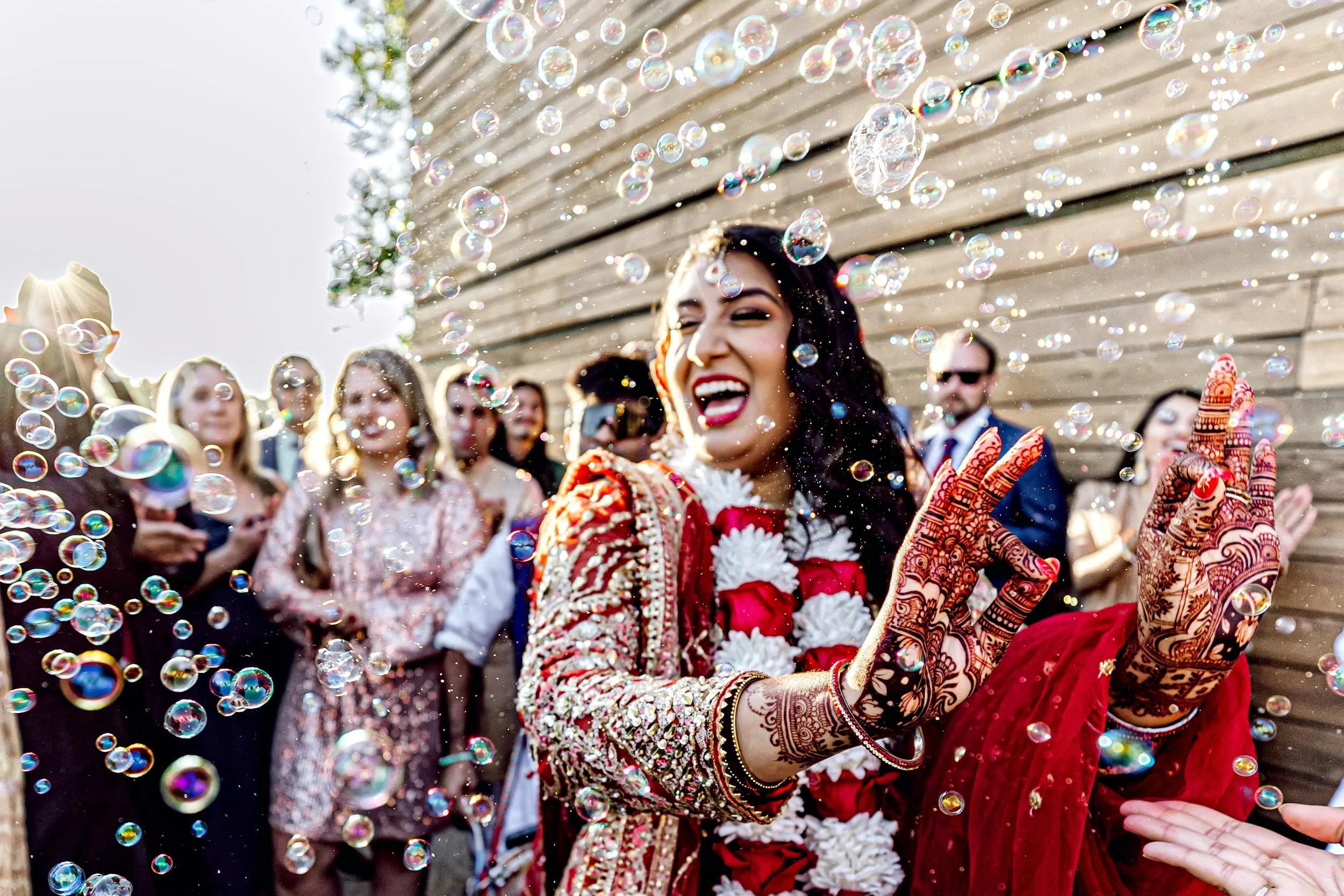 A woman in traditional Indian attire smiling and playing with soap bubbles at a celebration, surrounded by a group of people.