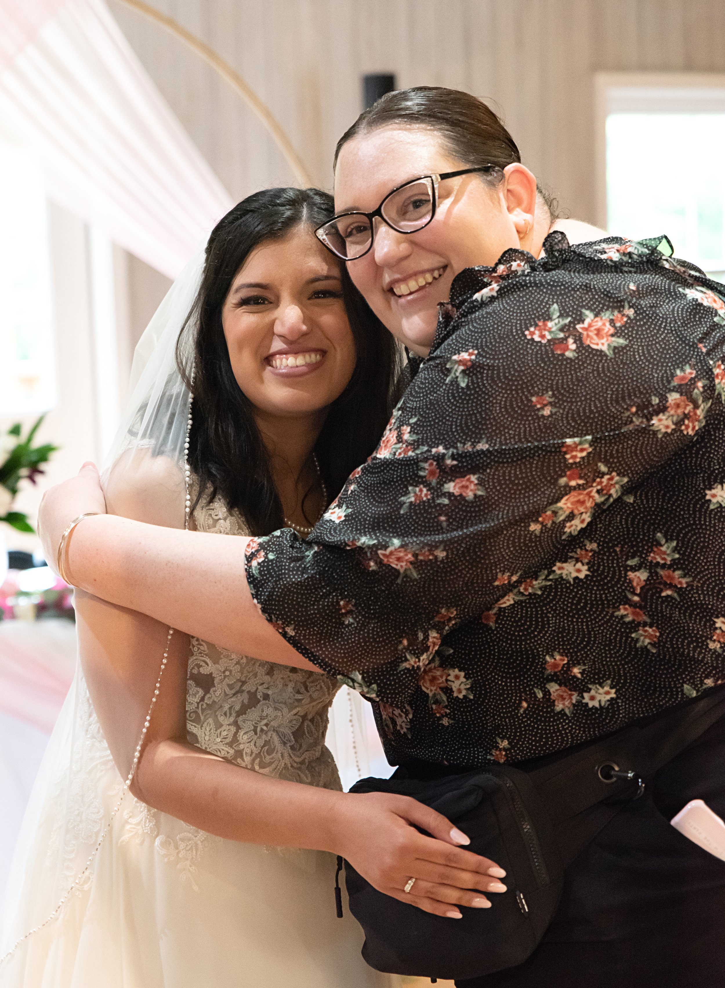 Two women hugging and smiling at a wedding. One woman is wearing a wedding dress and veil, and the other is wearing glasses and a floral shirt.