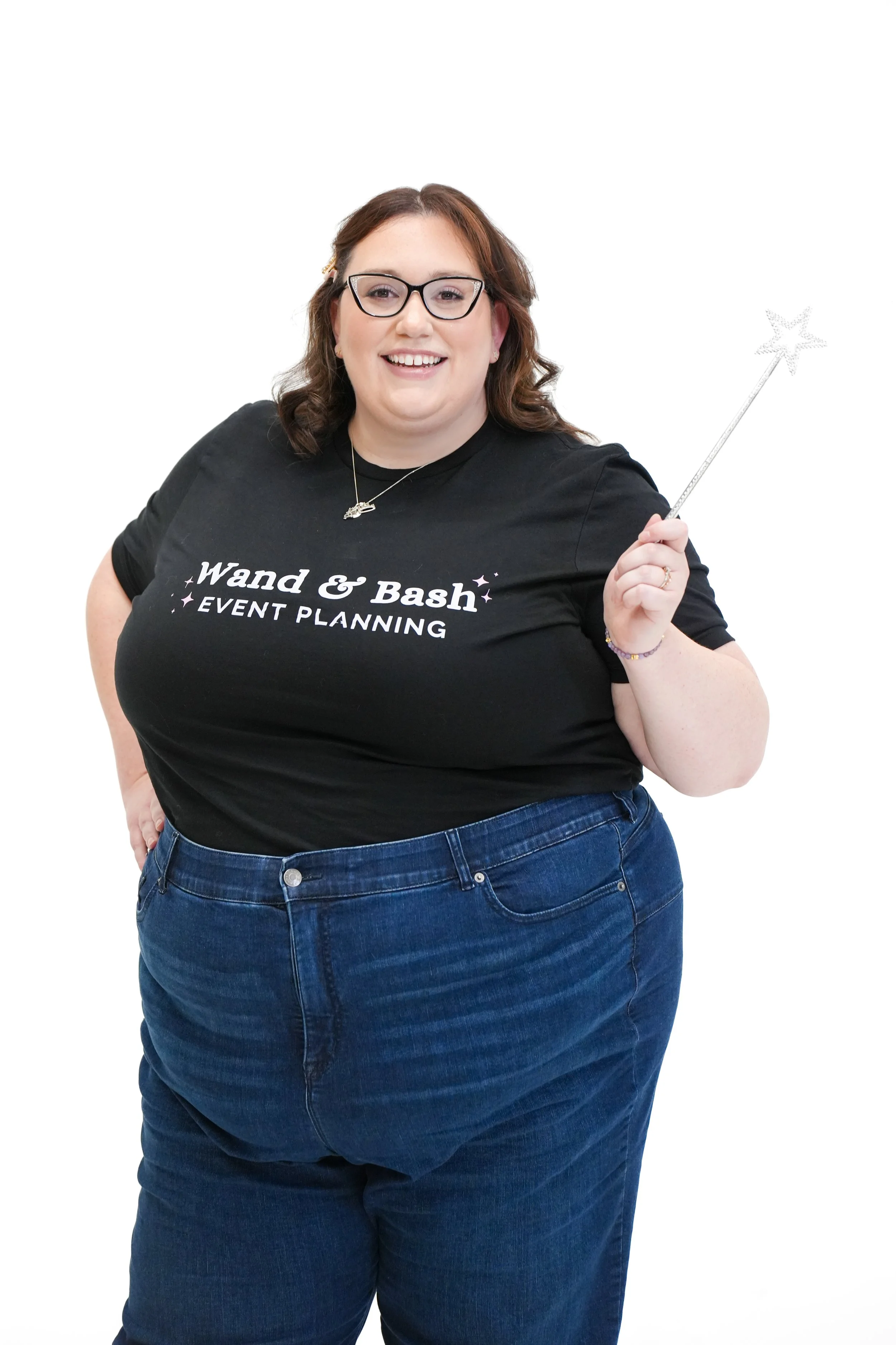 A smiling woman with glasses and brown hair, wearing a black T-shirt that says "Wand & Bash Event Planning" and blue jeans, holding a magic wand.