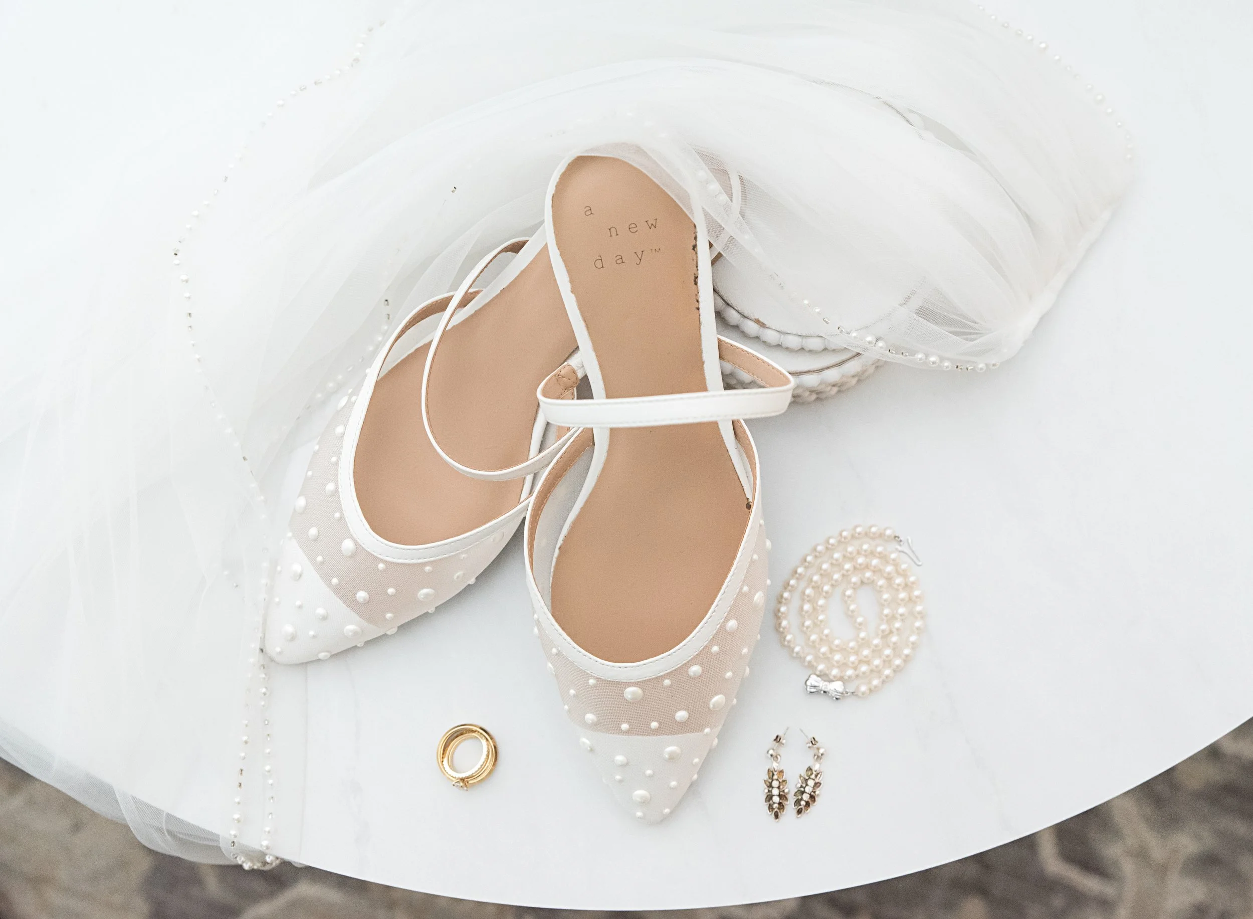 White bridal shoes with pearls, a pearl necklace, earrings, and a gold ring with a pearl setting on a white surface
