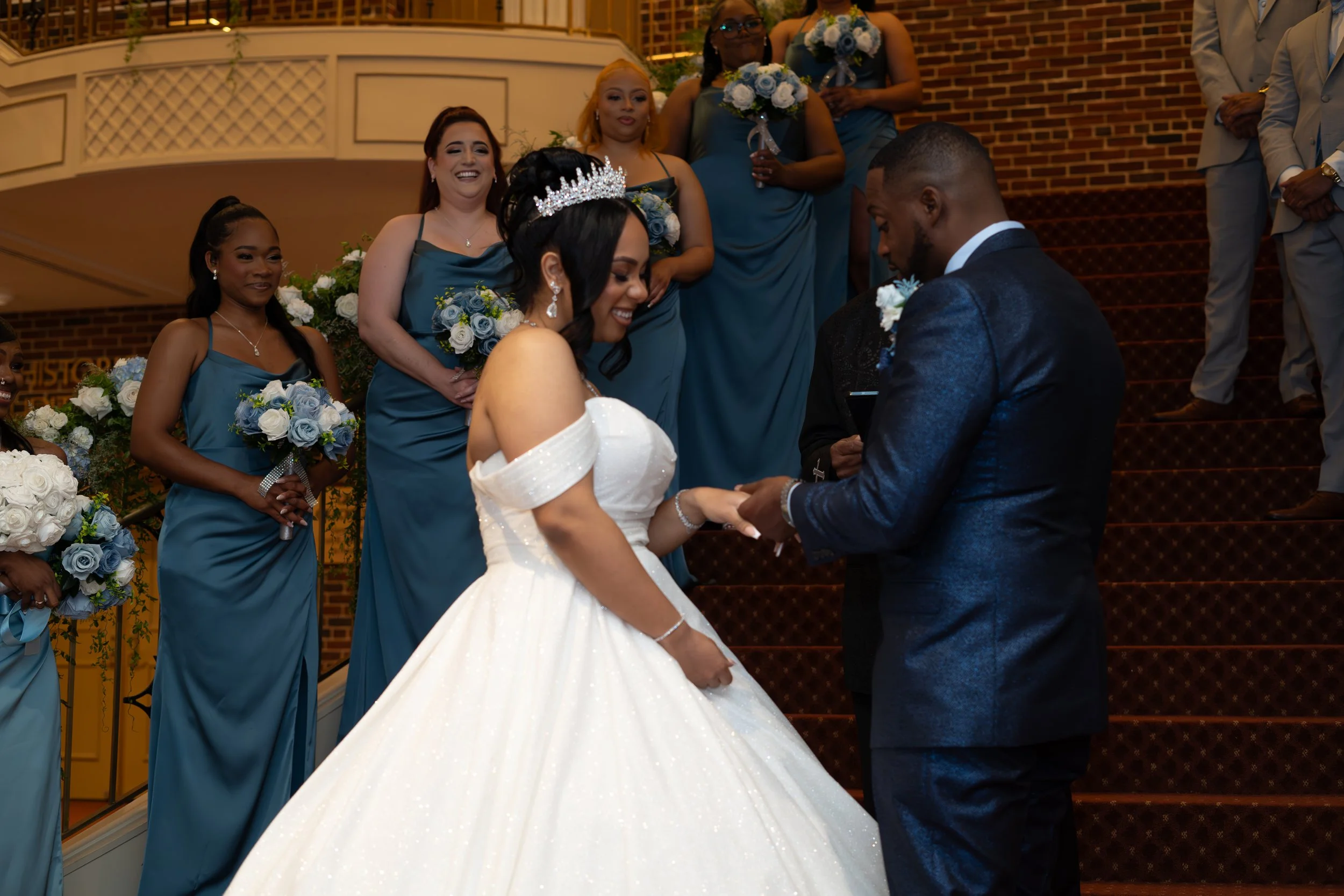 A bride and groom exchanging wedding vows on stairs, surrounded by bridesmaids and groomsmen, with the bridesmaids holding bouquets, during a wedding ceremony.
