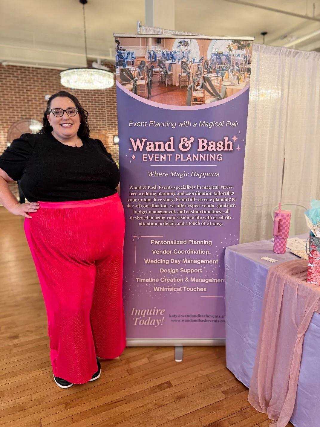 Woman in black top and bright pink pants standing next to a purple event planning banner at a wedding or event expo.