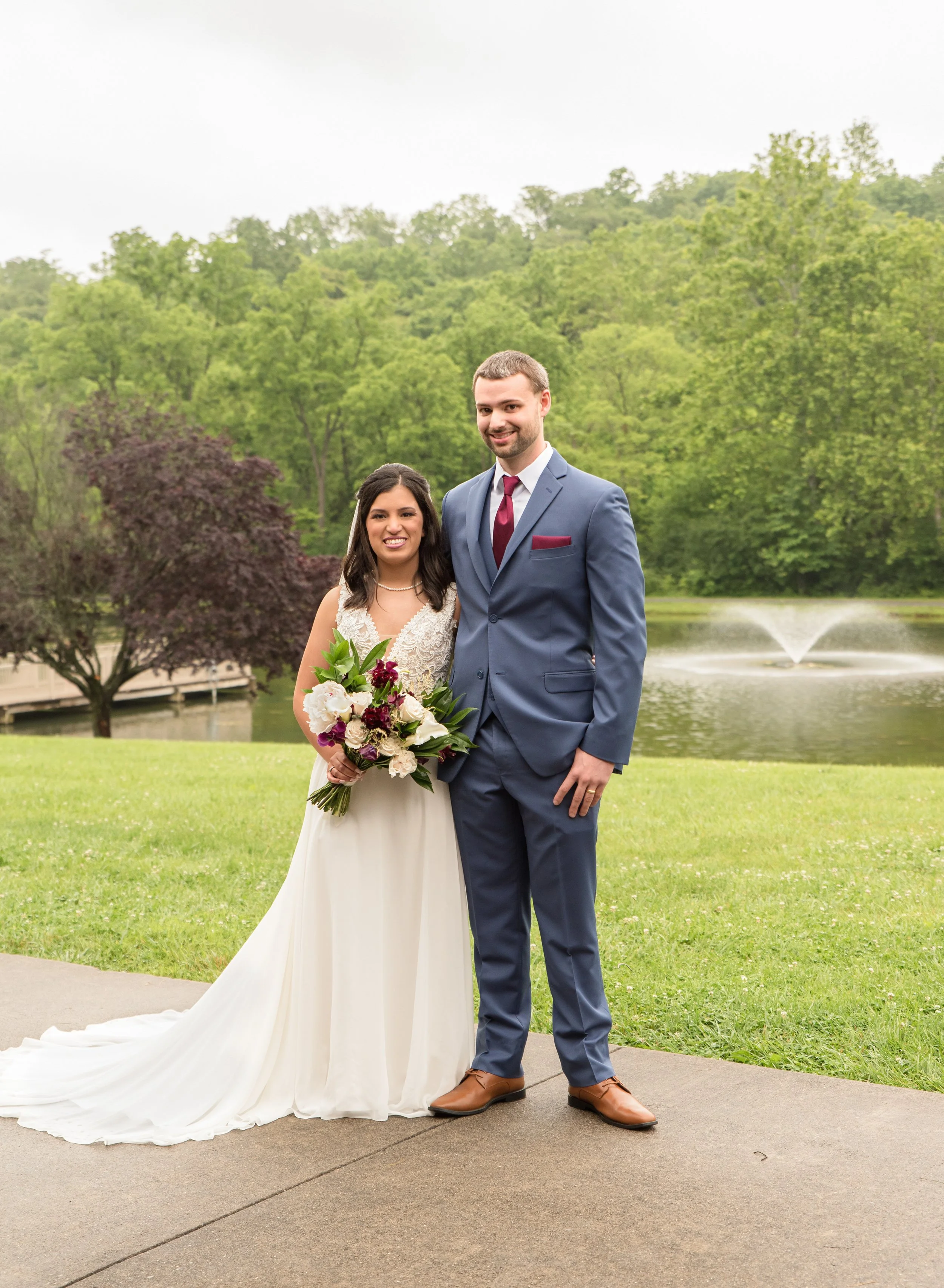 A bride and groom standing outdoors near a pond with a fountain, surrounded by green trees, on their wedding day.