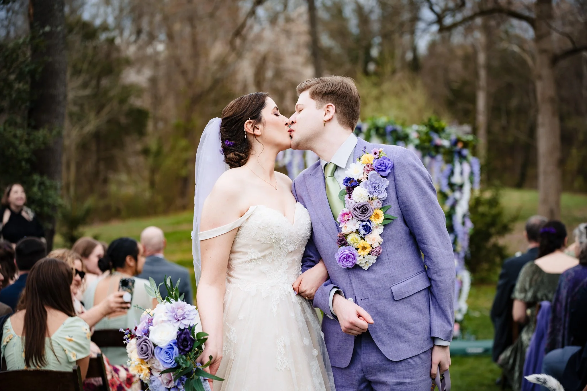 A bride and groom kiss during a wedding ceremony outdoors, surrounded by guests and trees.