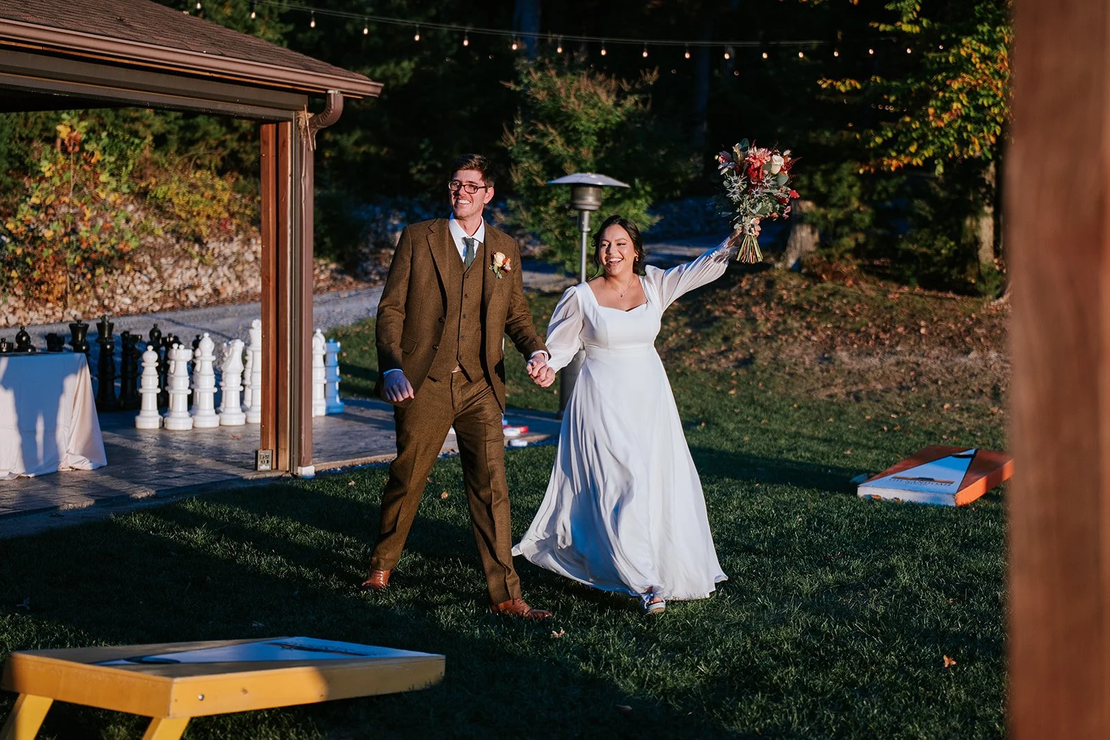 A newlywed couple holding hands, smiling, and celebrating outdoors during their wedding reception, with the bride holding a bouquet and dressed in a white gown, and the groom in a brown suit.