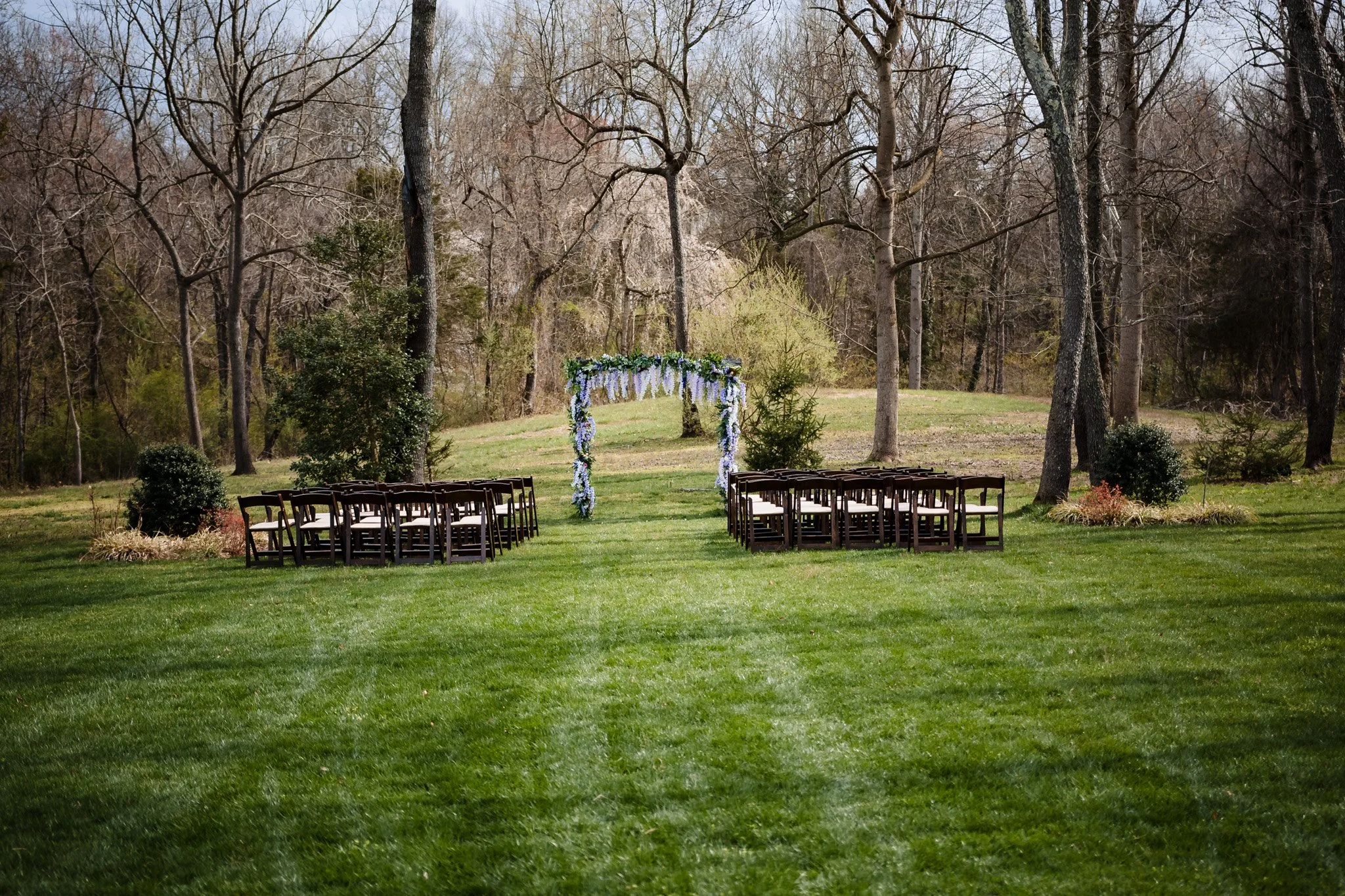 Outdoor wedding ceremony setup in a grassy area surrounded by trees, with chairs arranged in rows on either side of a floral decorated arch.