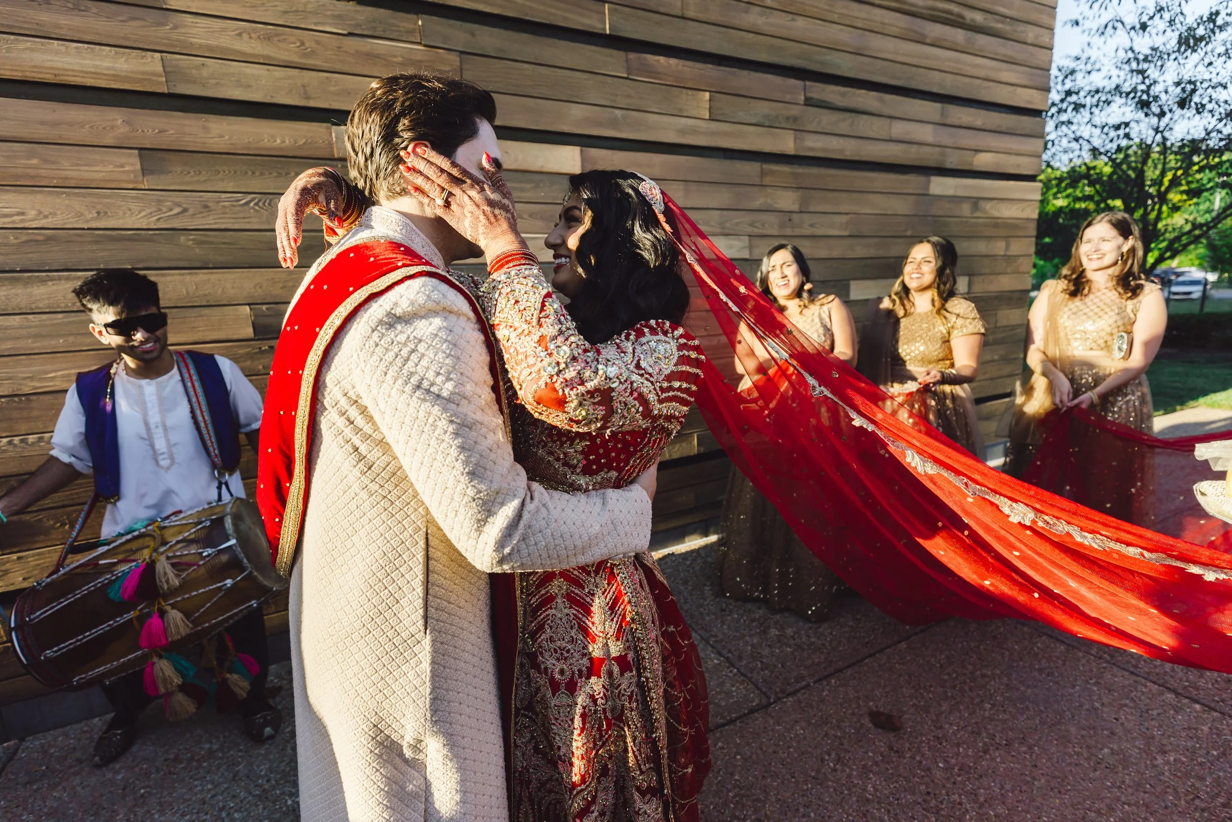 A wedding couple with the bride in a red embroidered dress and veil, and the groom in a cream traditional outfit, sharing an intimate moment outdoors with friends in gold dresses and a musician playing drums in the background.