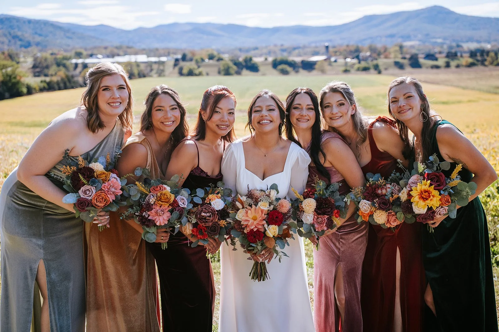 Group of women in formal dresses holding bouquets of flowers, standing outdoors with mountains and fields in the background.
