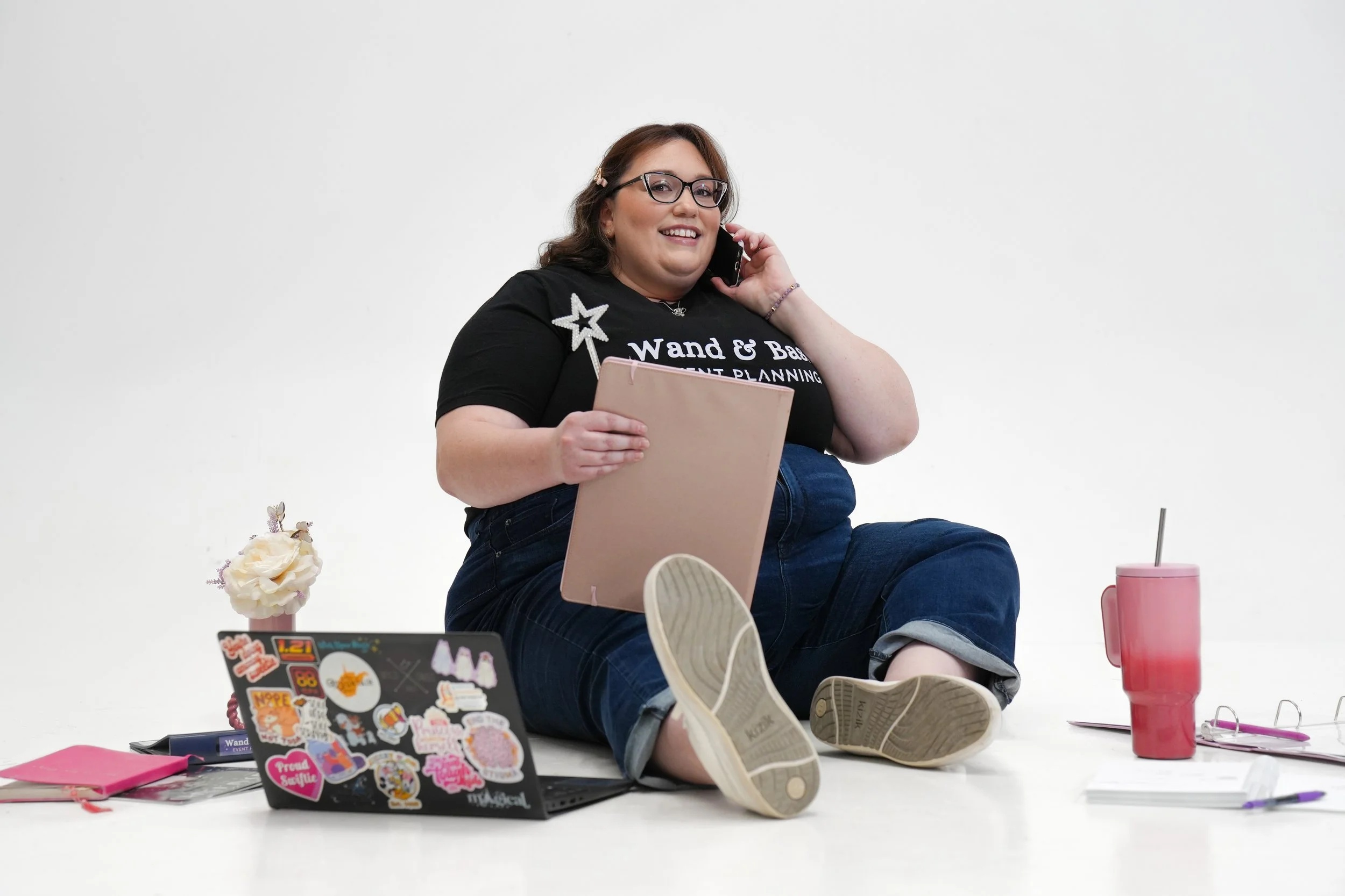 Person sitting on the floor, smiling, talking on the phone, surrounded by a laptop with stickers, notebooks, a pink tumbler, and decorative flowers against a plain white background.
