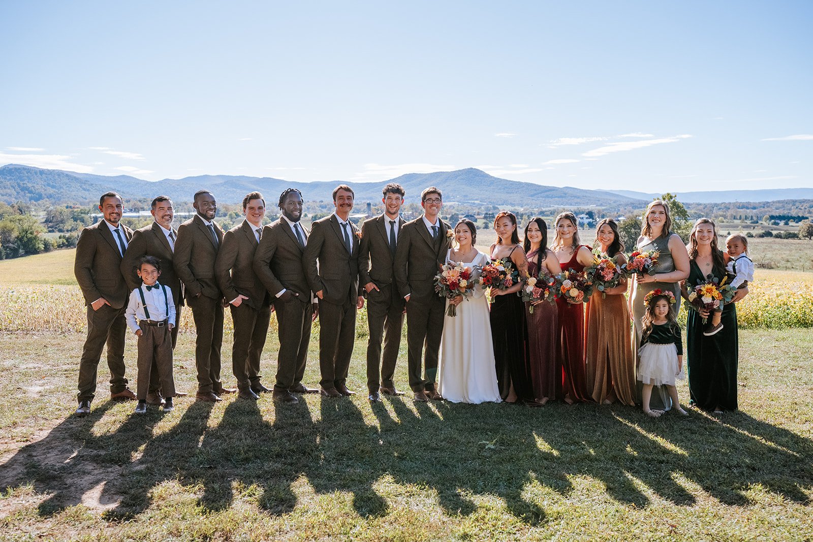 Group of wedding party members standing outdoors in a scenic landscape with mountains in the background, dressed in formal attire and holding bouquets.