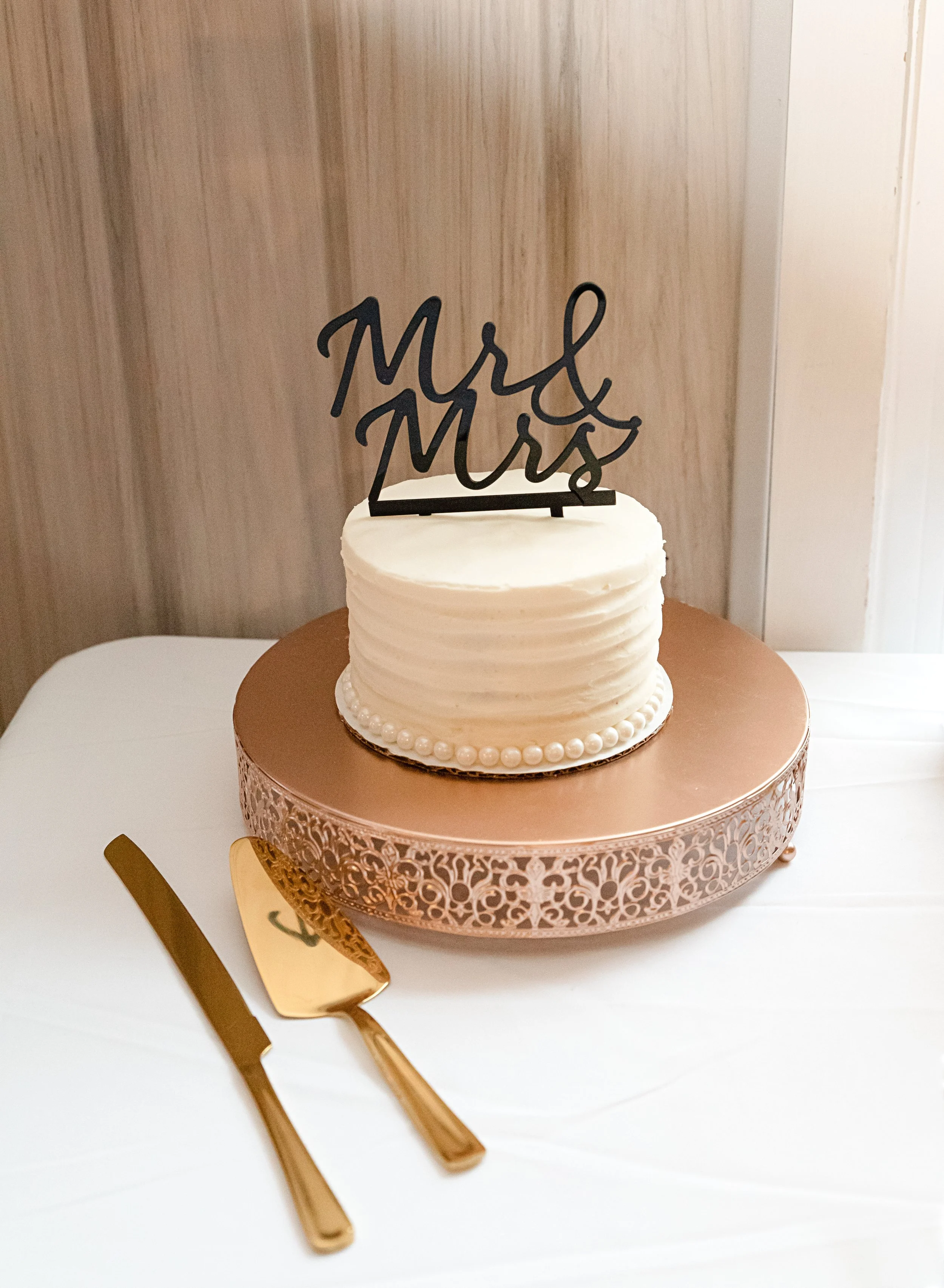 A wedding cake with white icing and pearl decoration, topped with a black "Mr & Mrs" cake topper, is on a decorative pink cake stand. There is a gold cake knife and server on the table in front of the cake.