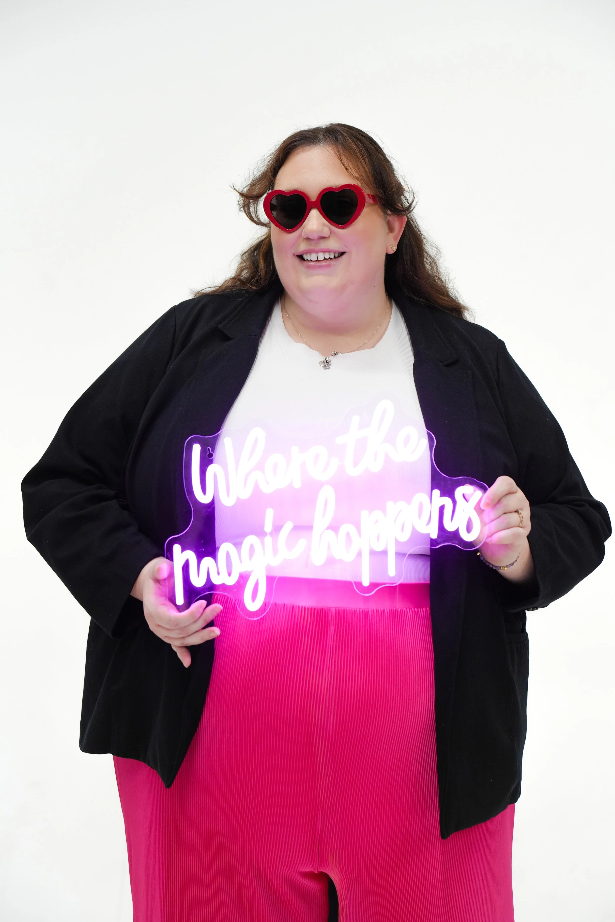 A smiling woman wearing heart-shaped sunglasses, a black jacket, a graphic white t-shirt, and pink pants, holding a purple neon sign with white cursive text that reads 'where the magic happens', against a plain white background.