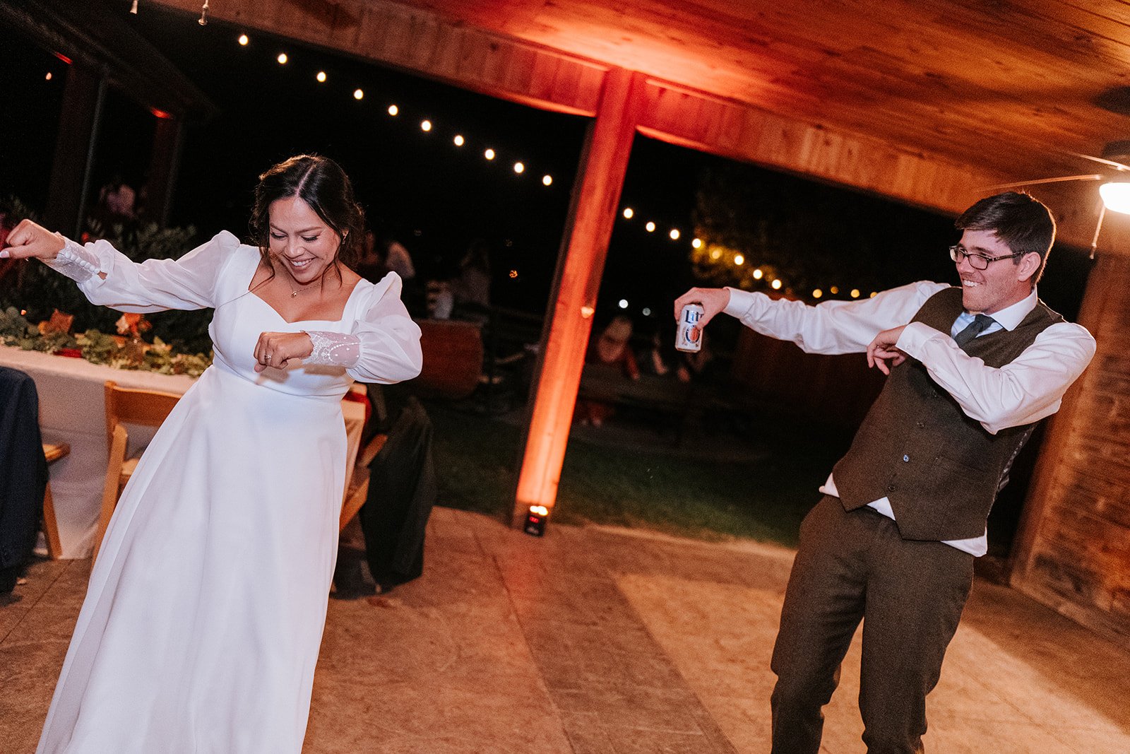 A woman in a white dress and a man in a brown vest and pants dancing at a nighttime gathering with string lights, with the woman smiling and the man holding a can of beer.