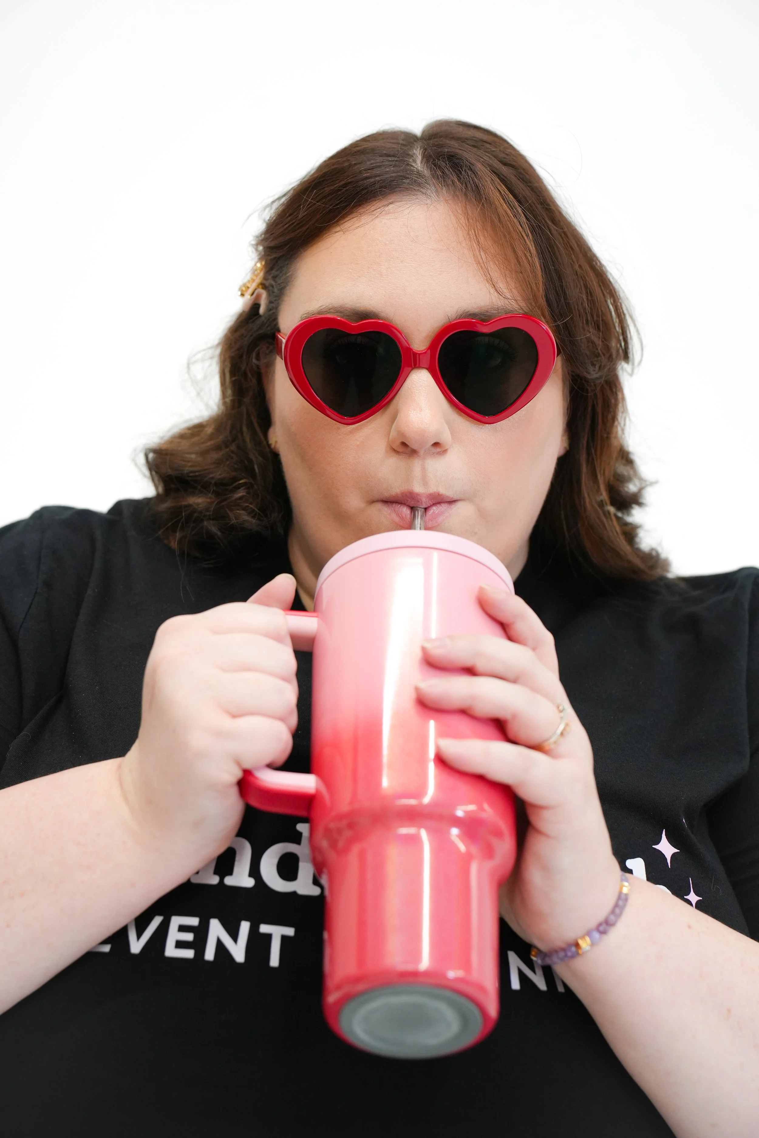 Woman with brown hair wearing heart-shaped sunglasses, drinking from a pink mug with a straw.