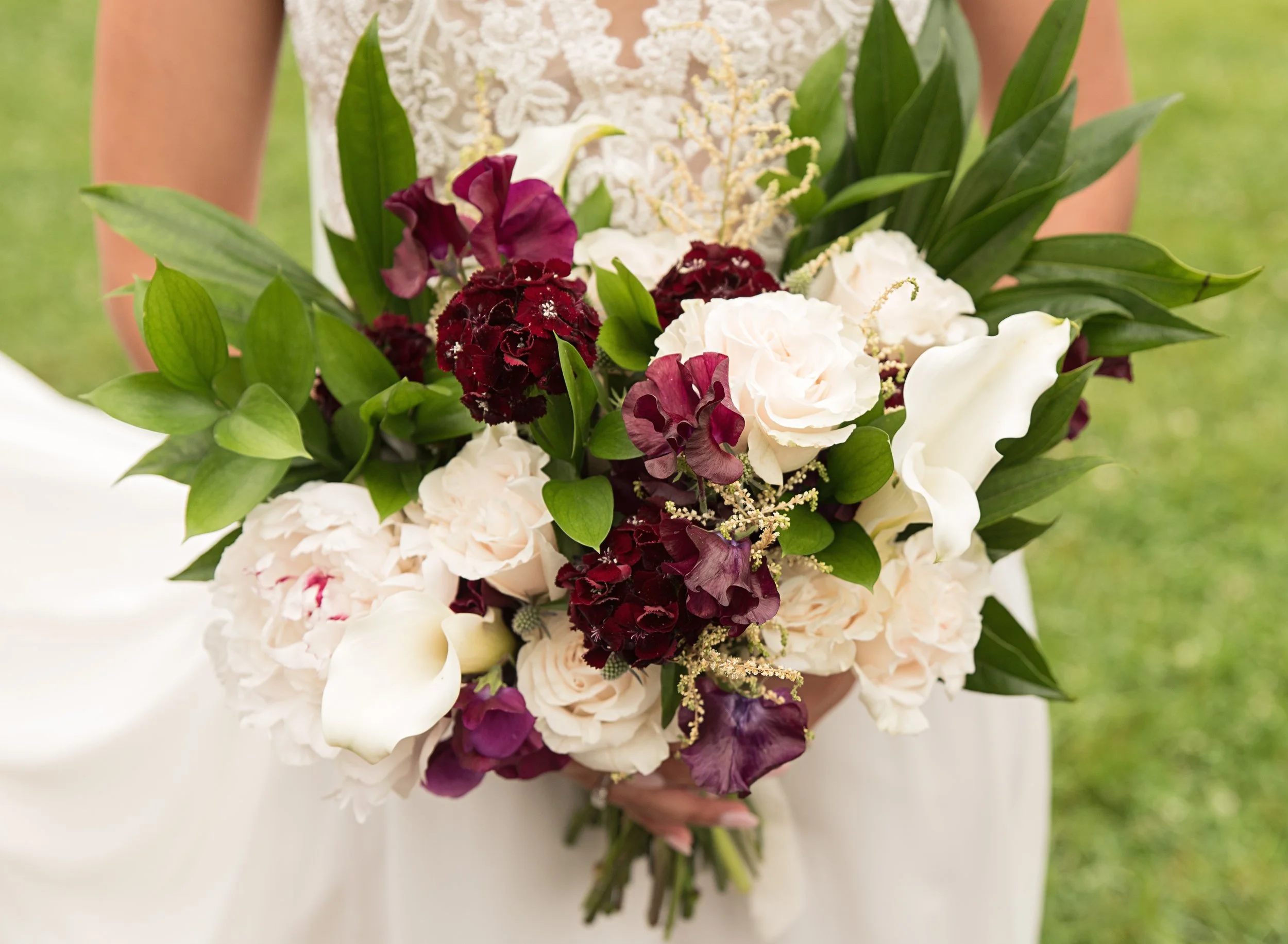 A woman holding a bouquet of white and deep burgundy flowers with green leaves, outdoors on grass.