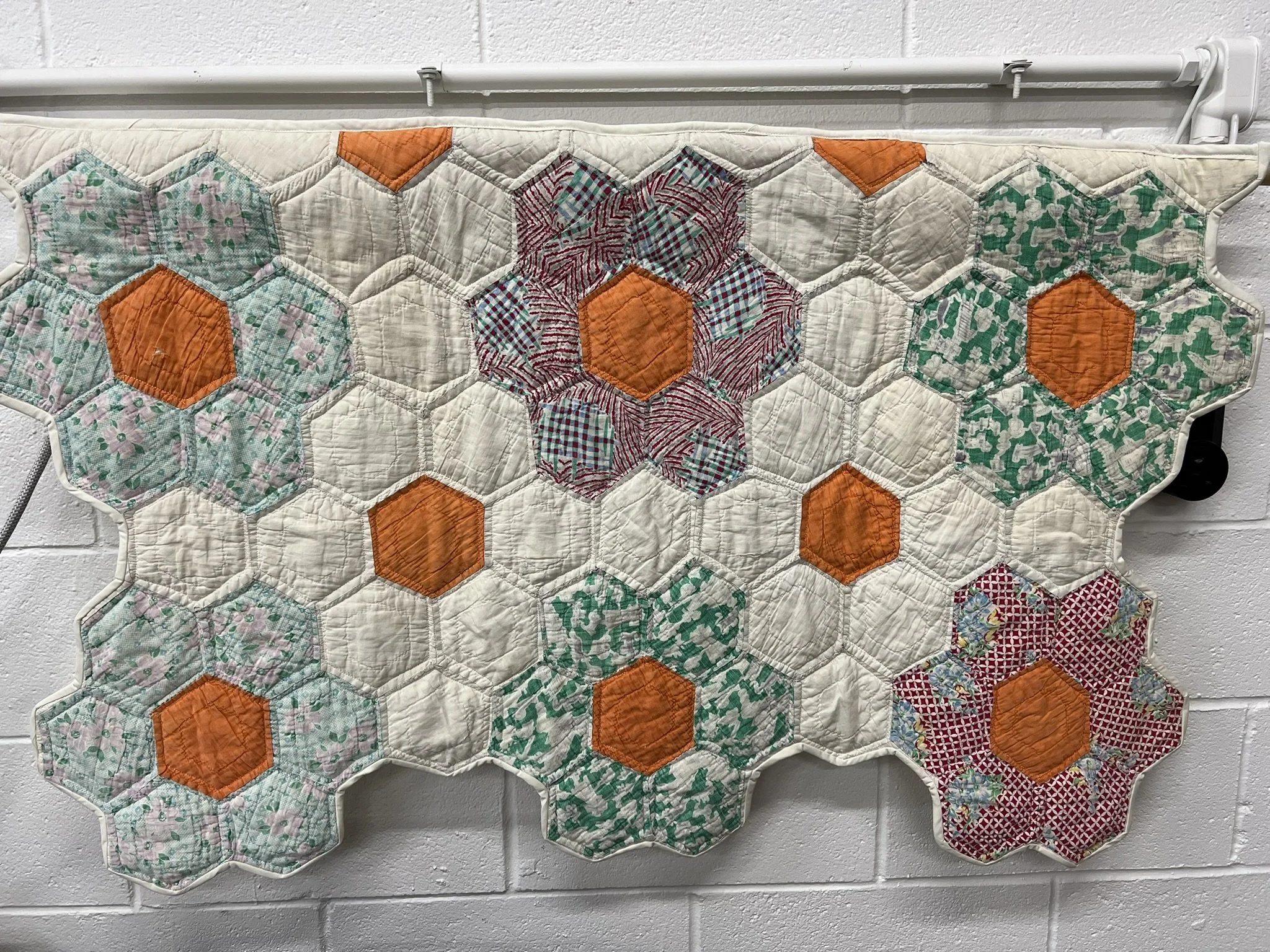 A quilt with hexagon and flower shapes in various colors and patterns, including orange, red, green, pink, and white, hanging on a wall with a white background.