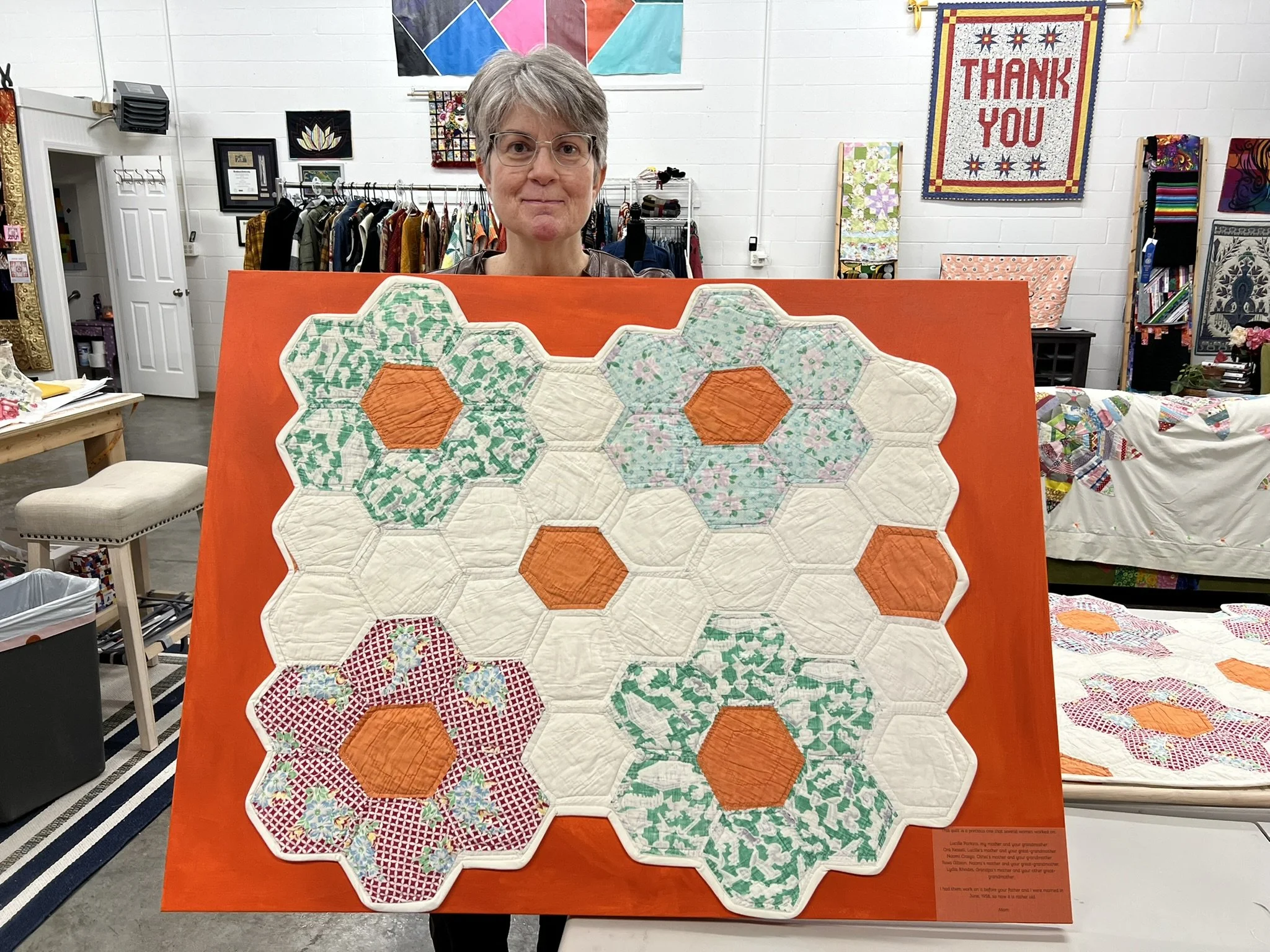 Woman holding a quilt with honeycomb pattern, featuring hexagons with various colorful fabrics, in an indoor craft room.