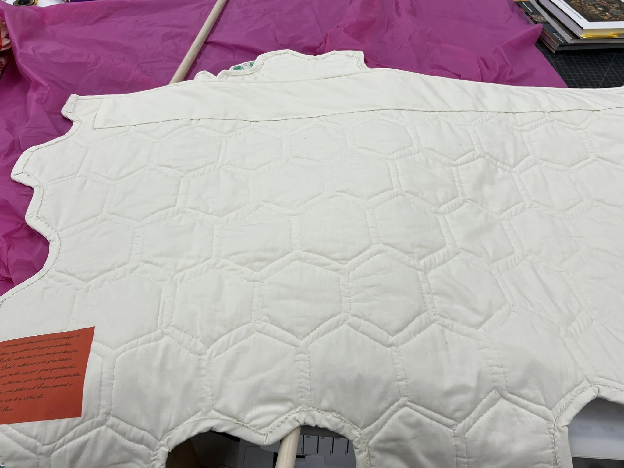 A white quilt or padded fabric with a honeycomb pattern laid out on a table with pink fabric and books in the background.