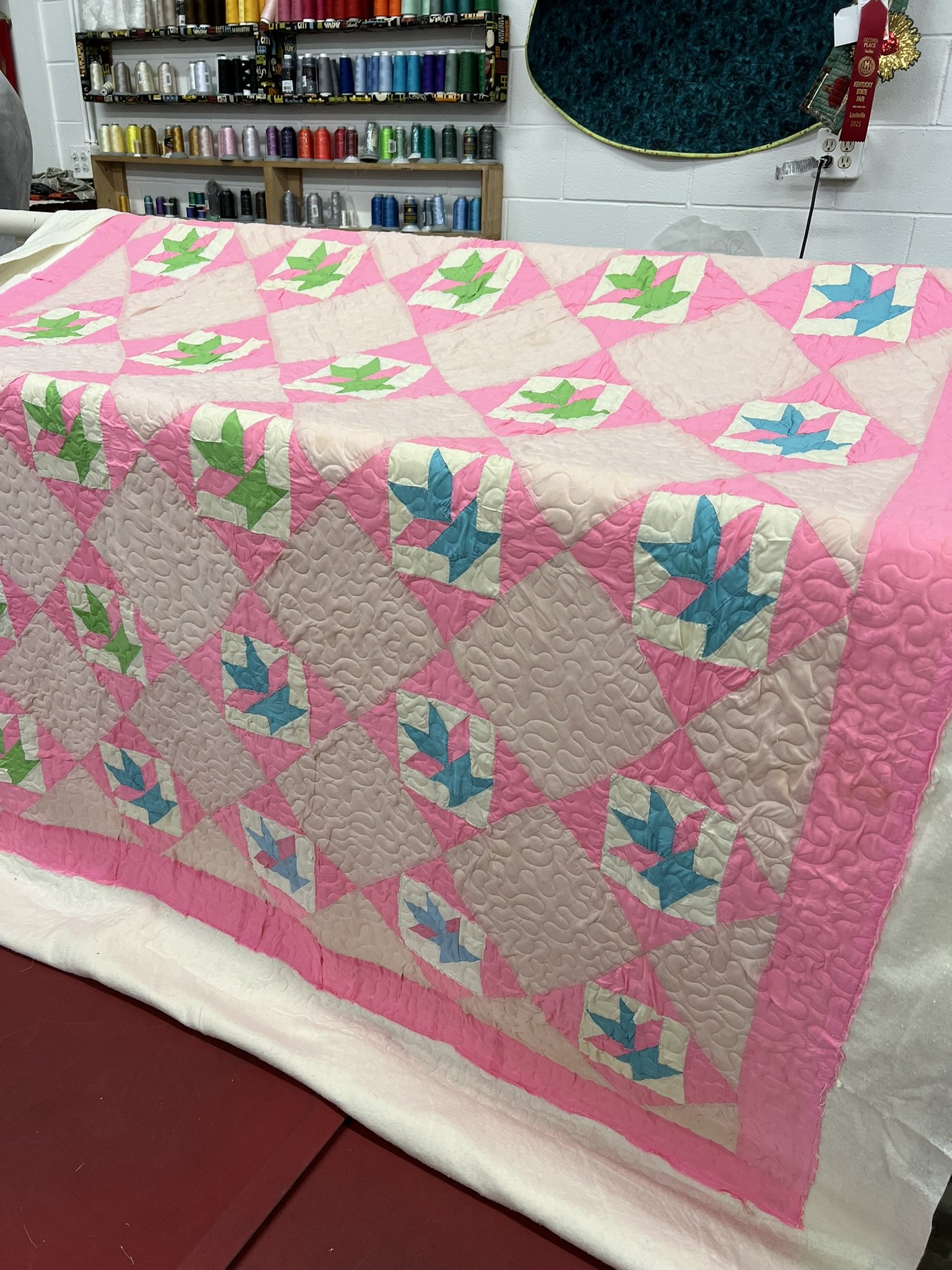 A colorful quilt with pink, beige, green, and blue geometric patterns draped over a surface, with shelves of thread spools and sewing supplies in the background.