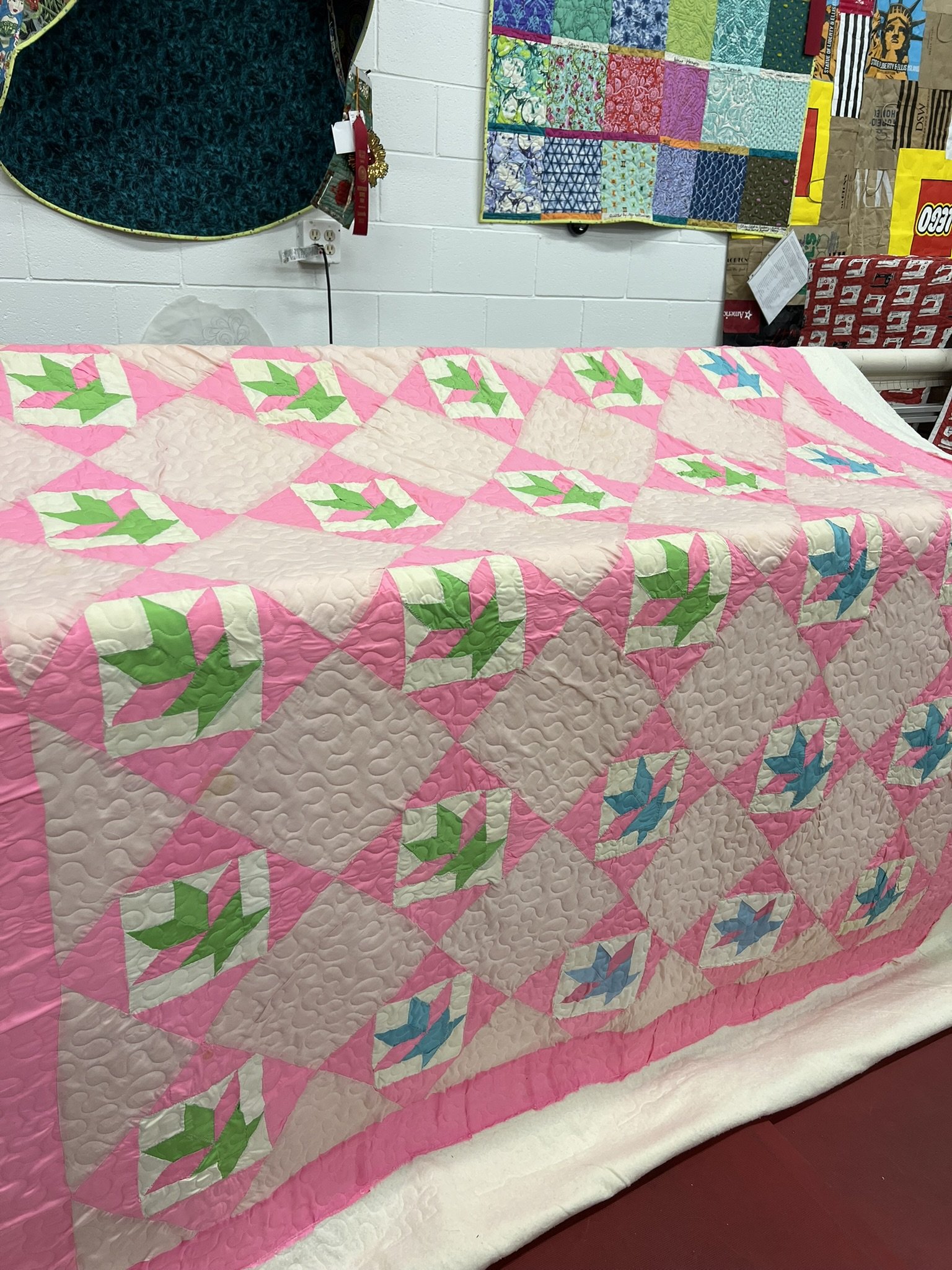 A colorful quilt with pink, beige, green, and blue patches featuring a holly leaf pattern, displayed on a bed in a quilting shop.