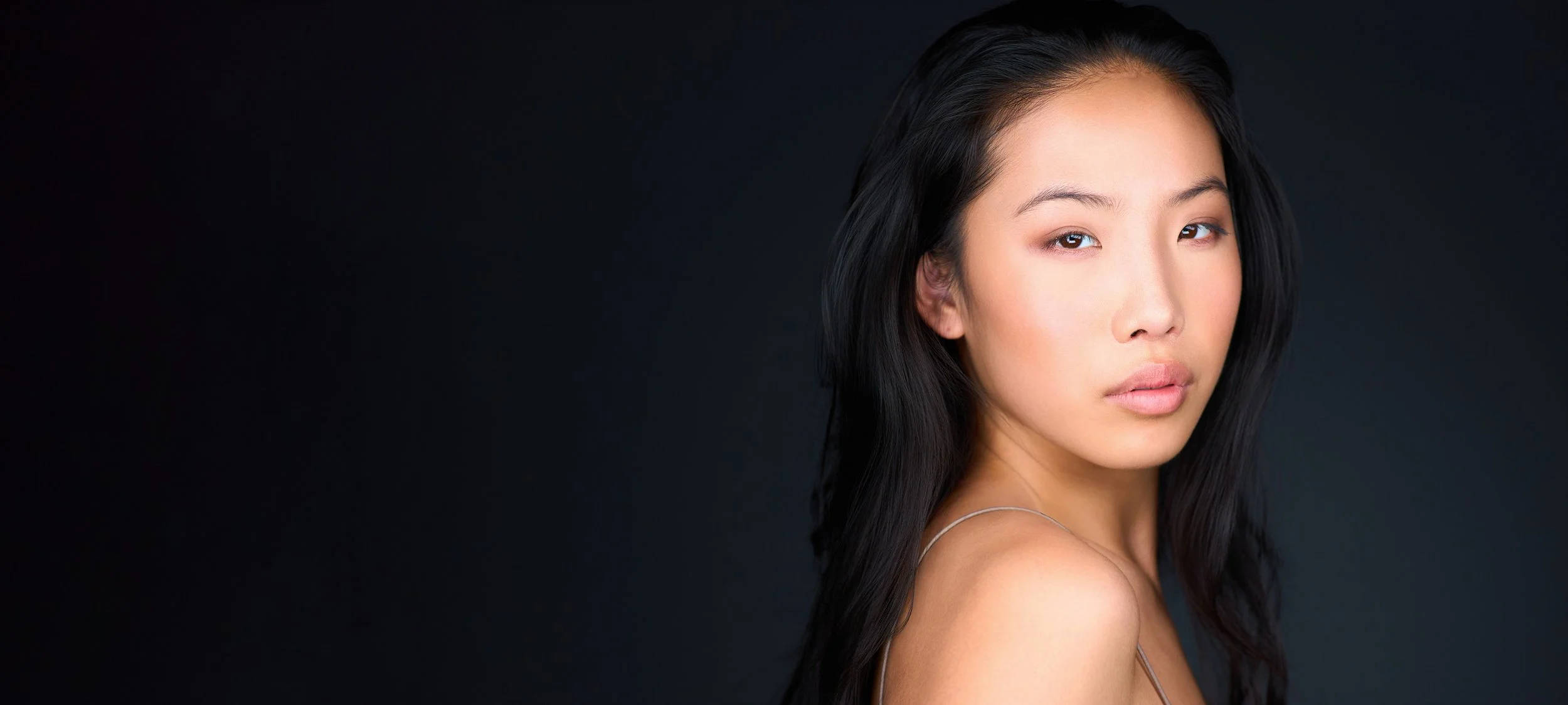 A headshot photo banner of a young woman with long black hair and natural makeup looks at the camera against a dark background.