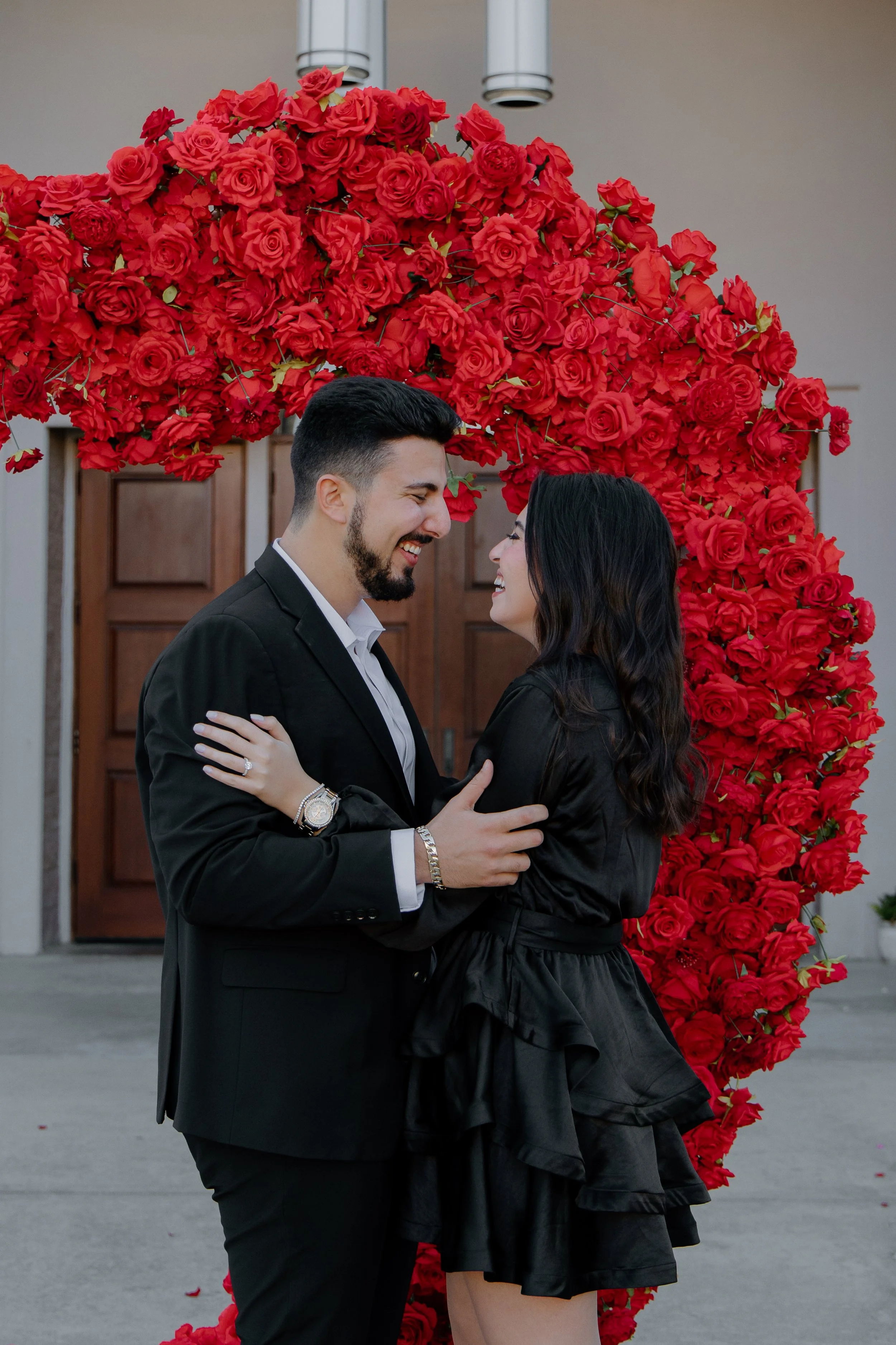 A romantic couple, dressed in black, sharing a happy moment in front of a large heart-shaped floral arrangement made of red roses.