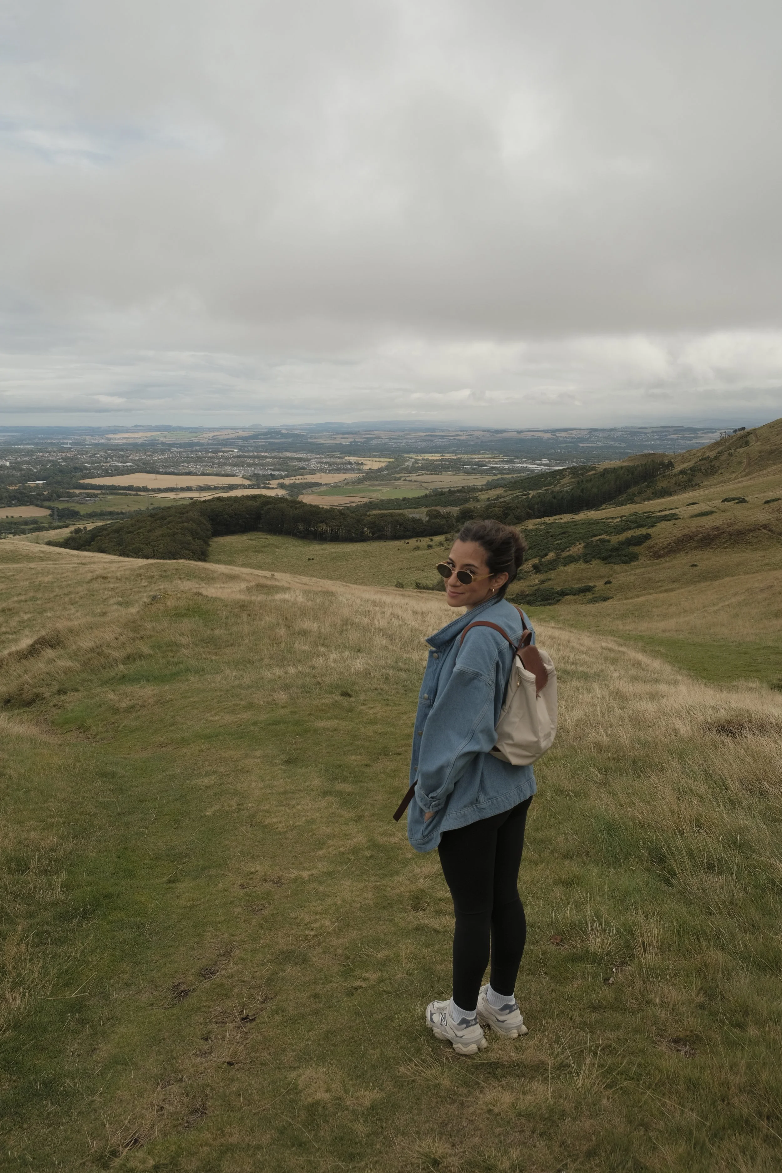 A woman standing on a grassy hillside with a backpack, wearing sunglasses, a denim jacket, leggings, and sneakers, overlooking a scenic, cloudy landscape with rolling hills and fields in the distance.