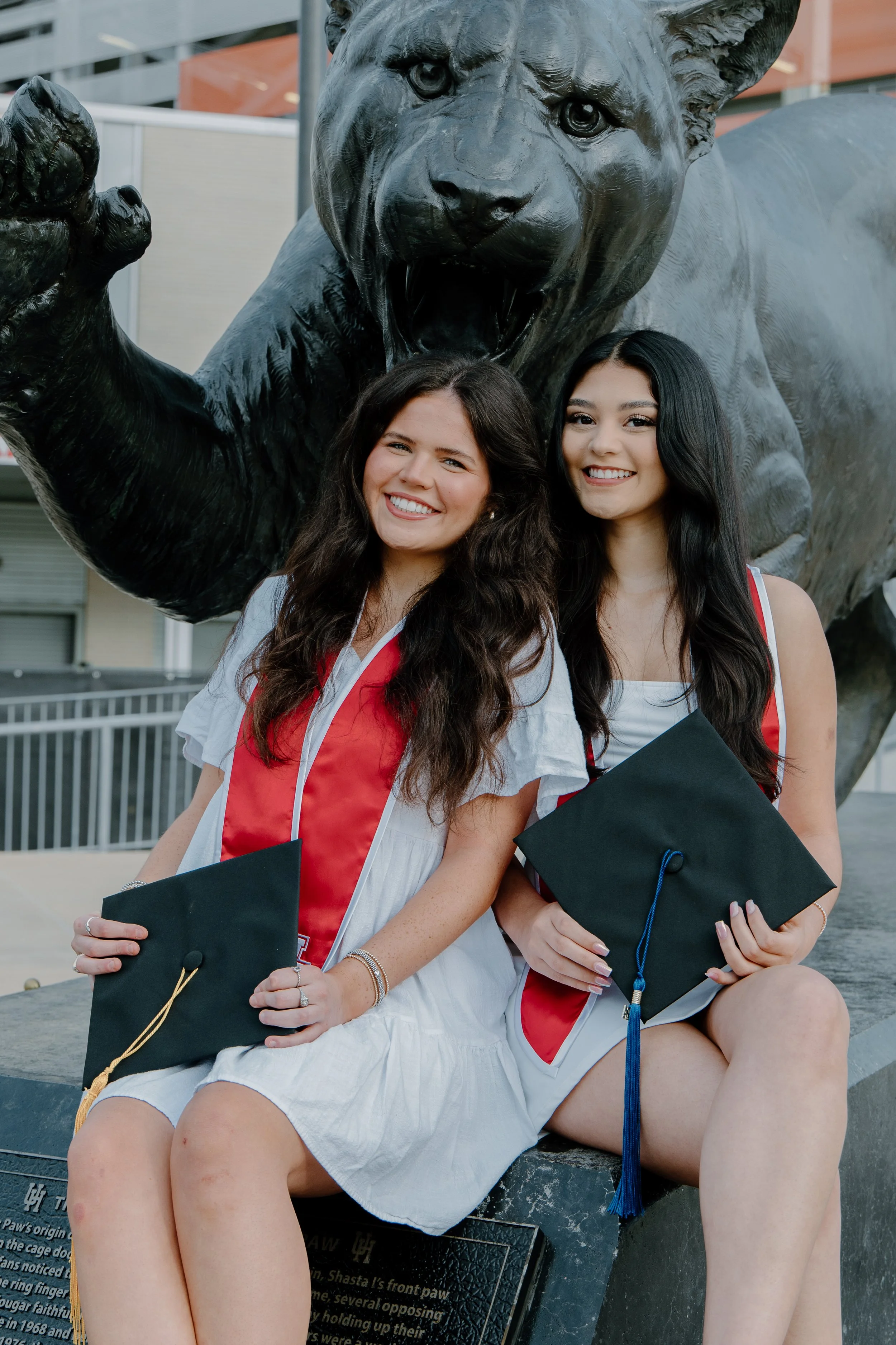 Two young women in graduation caps and gowns sitting in front of a large bear statue, smiling.