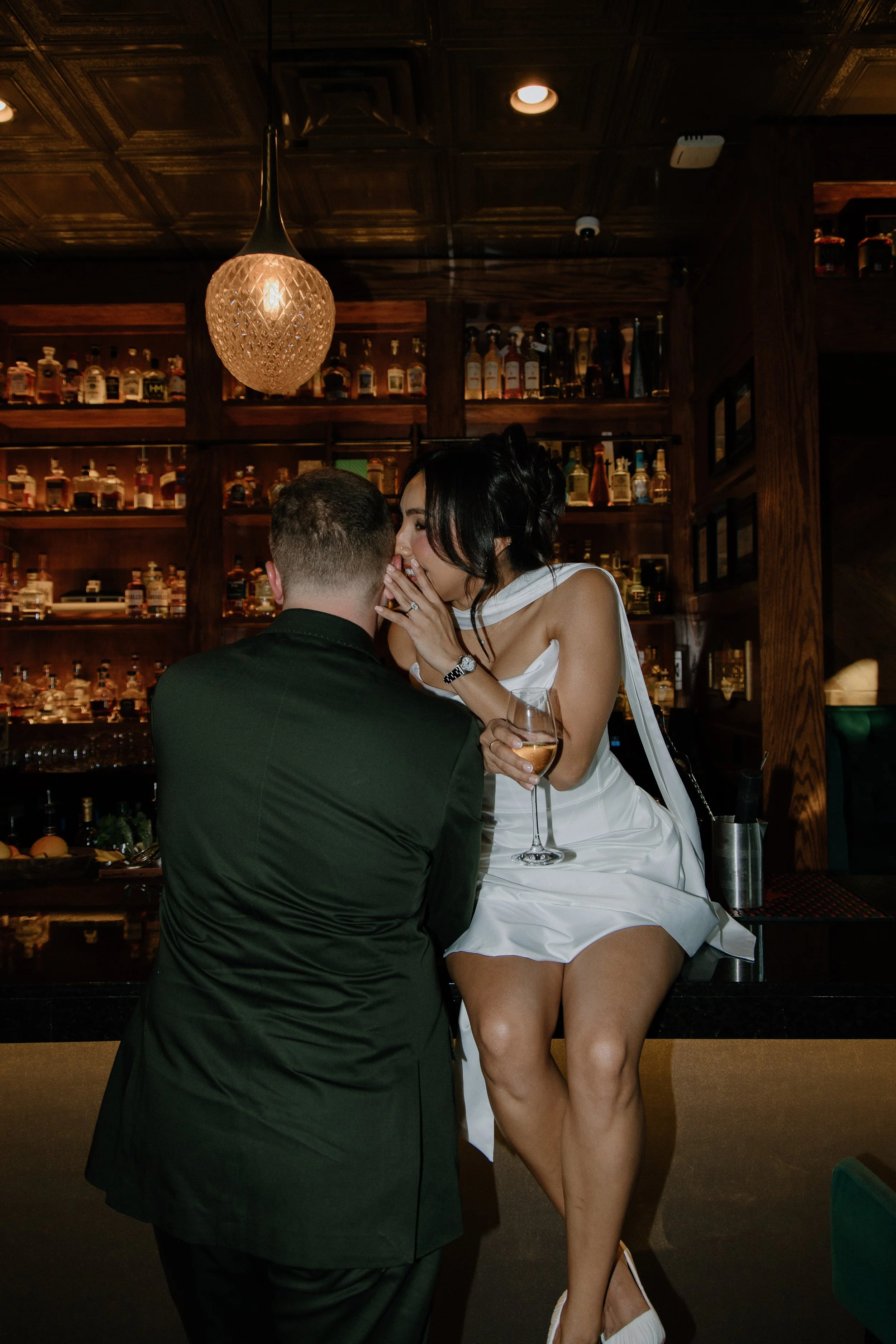 A woman in a white dress sitting on a bar counter, whispering into the ear of a man in a dark suit, while holding a glass of white wine, in a dimly lit bar with wooden shelves of liquor bottles in the background.