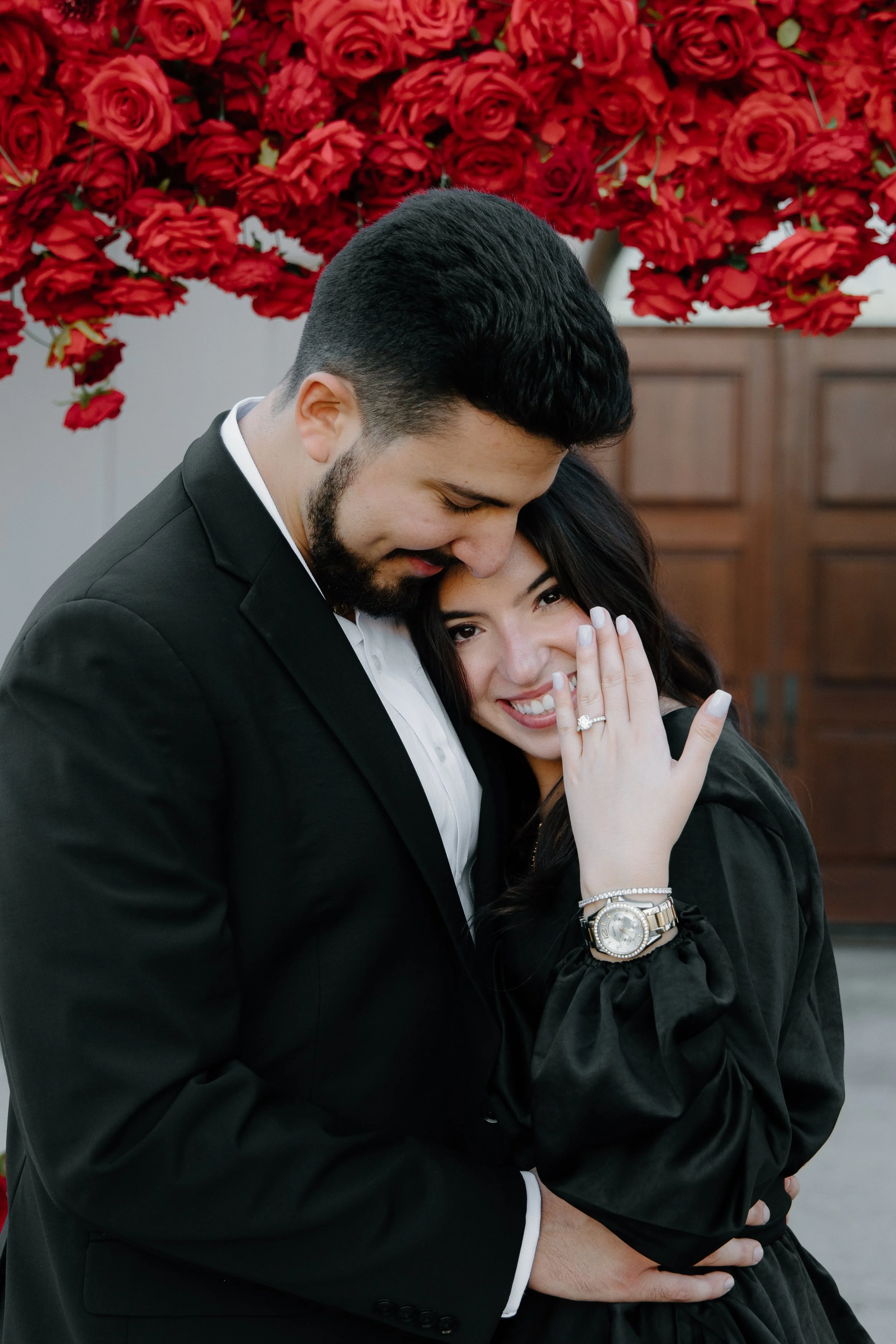 A couple embracing, the man in a black suit and the woman in a black dress, with her showing an engagement ring, standing in front of a backdrop of red roses.