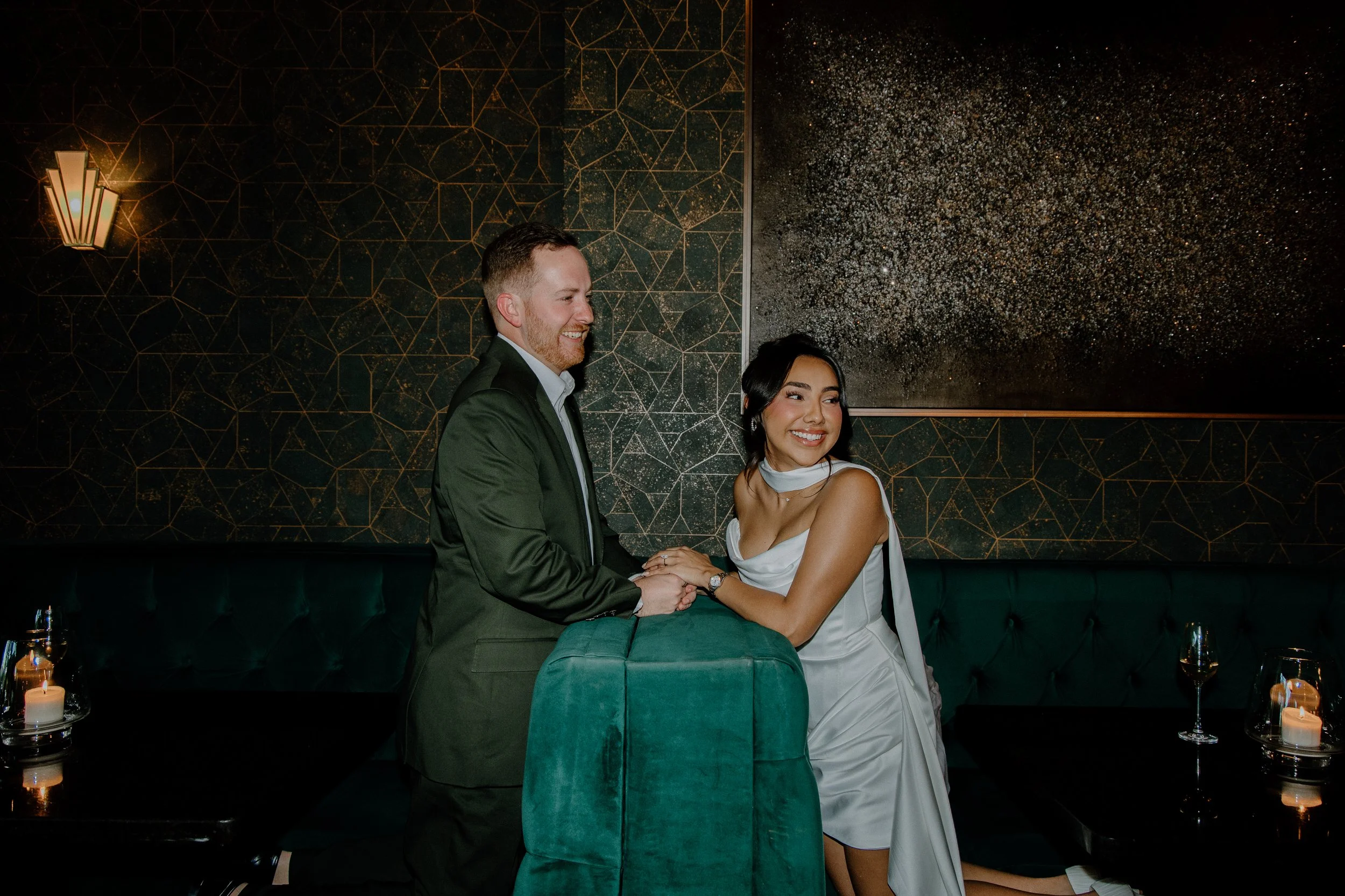 A happy couple holding hands and smiling at each other in a dimly lit, elegant lounge with dark green seating, candles, and modern decor.