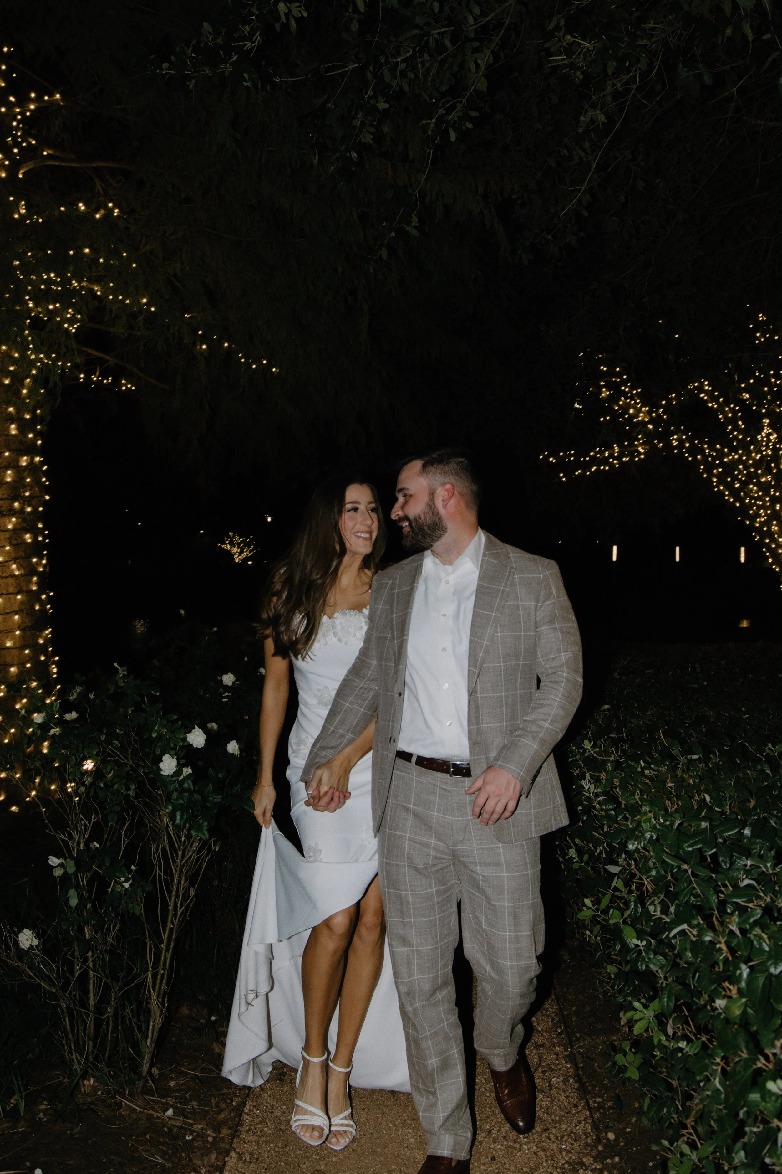 A couple in wedding attire walking hand in hand through a garden at night, surrounded by trees decorated with string lights.