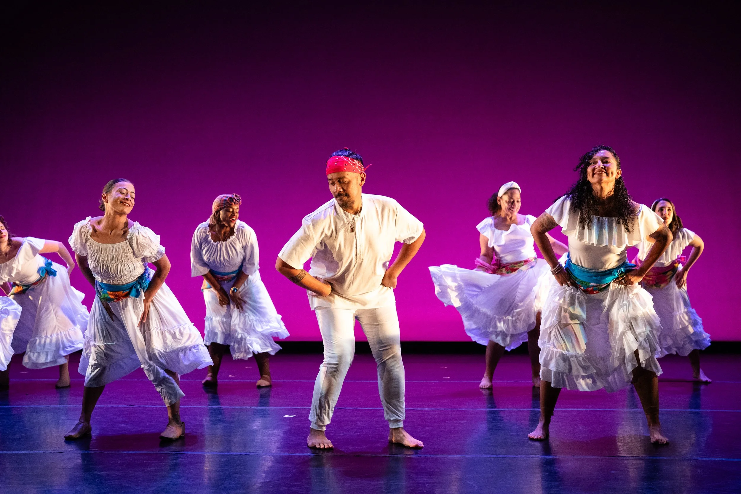 Group of dancers performing on stage with a purple background, dressed in traditional white costumes with colorful sashes, some wearing headscarves, and one male dancer wearing a red bandana, smiling and dancing.